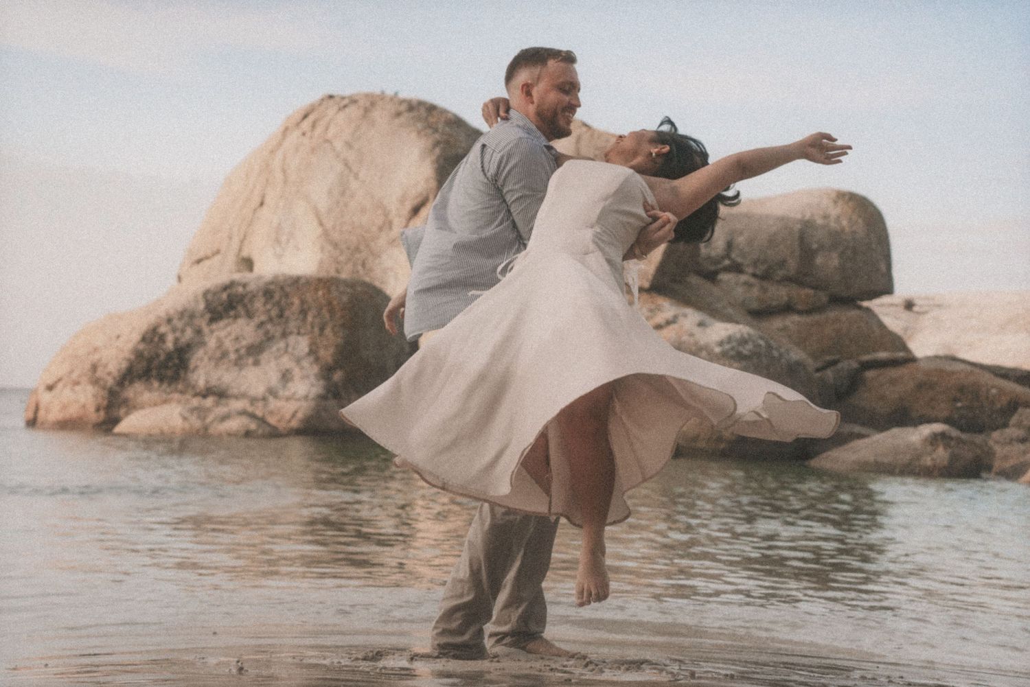 Couple dancing on the rocks in the shallows as her white dress lifts