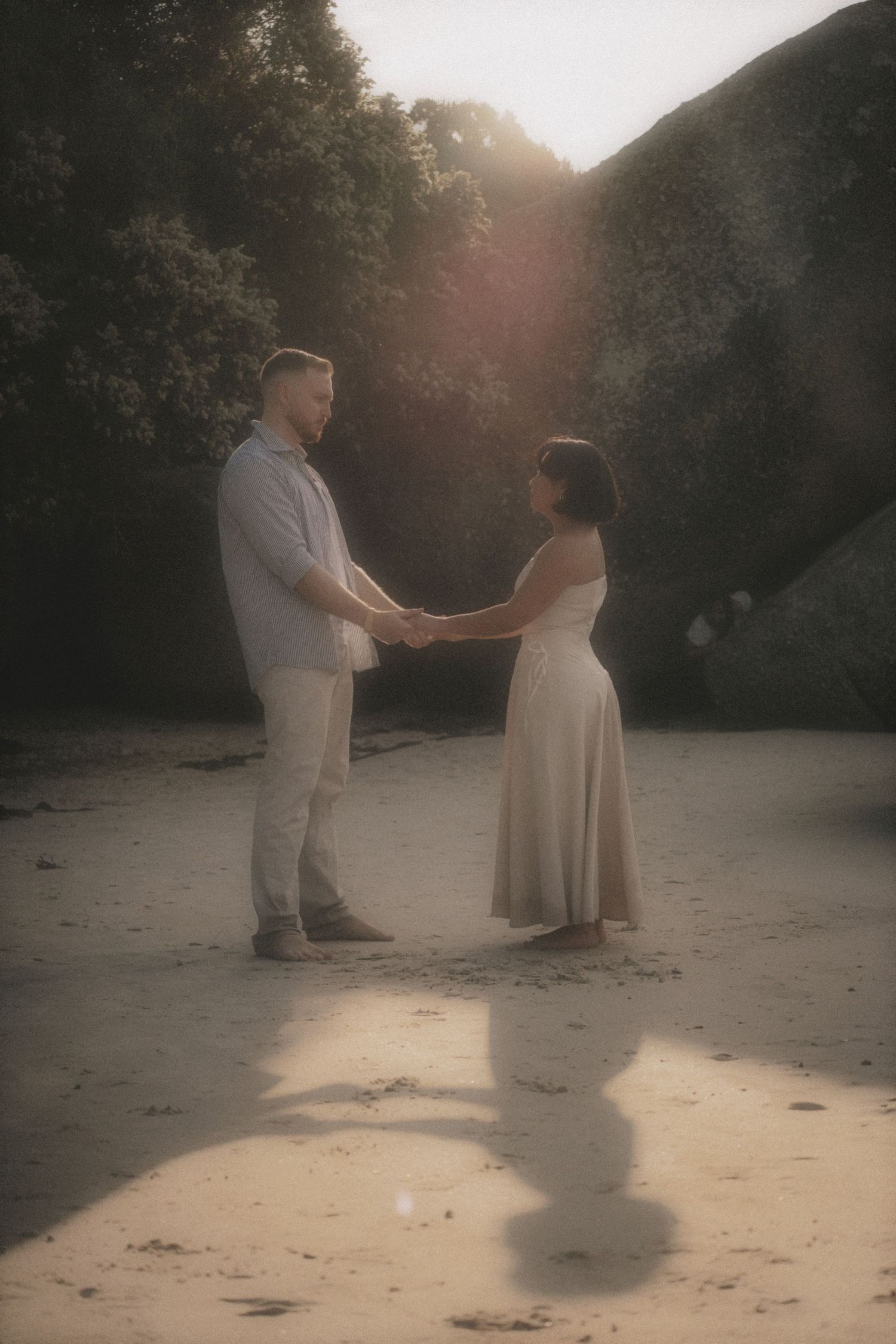 Couple holding hands and facing each other on a sandy cove, backlit with long shadows