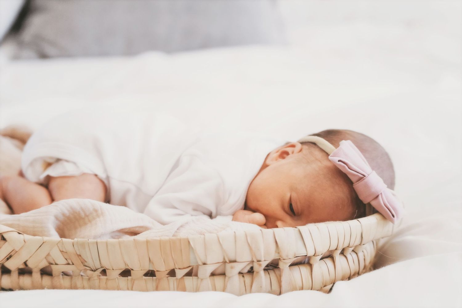 Sleeping newborn in a pink bow headband curled up in a woven basket on a bed