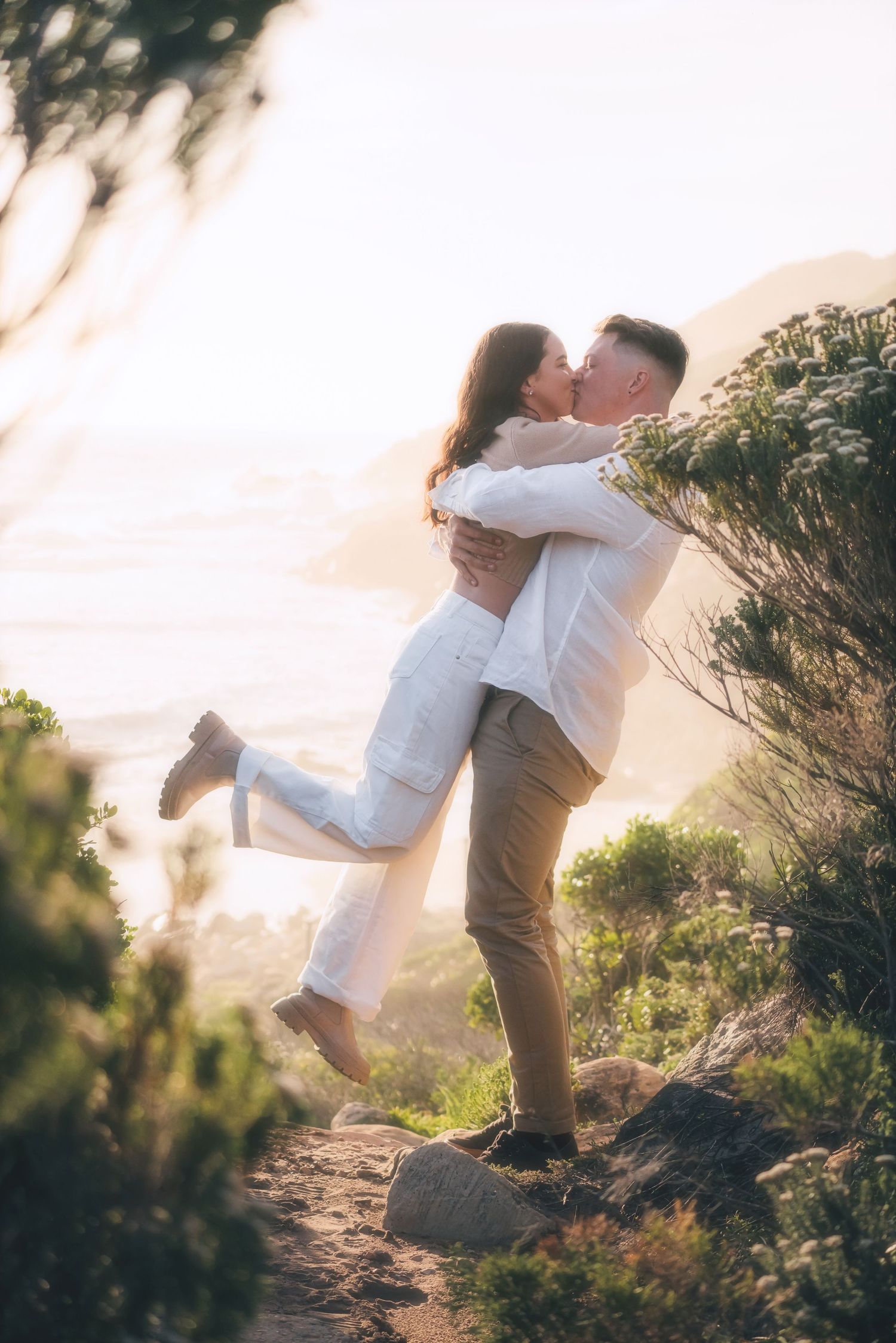 Couple kissing as he lifts her off the ground on a coastal path lined with fynbos