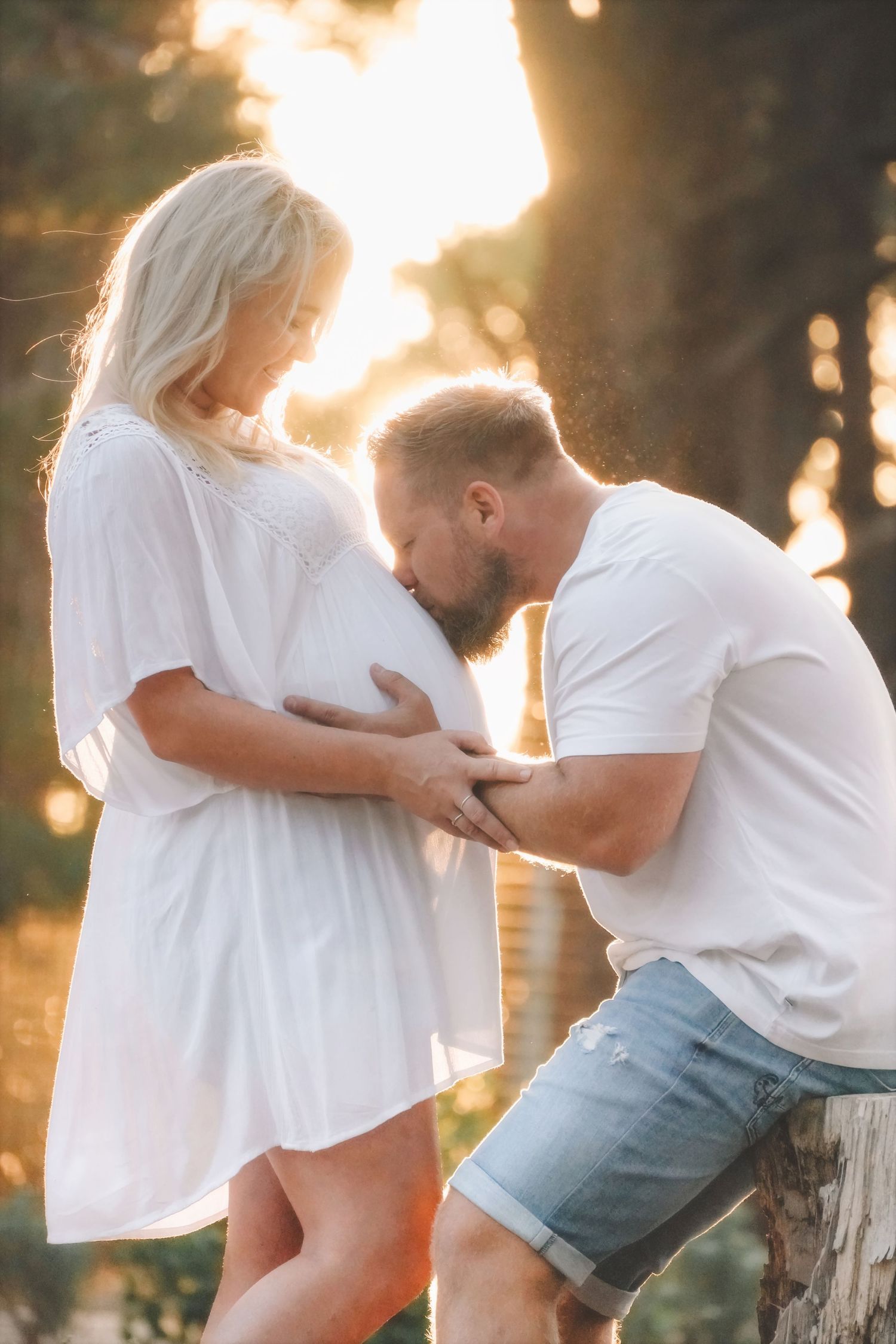 Man kissing his partner's baby bump in golden, sunlit forest light