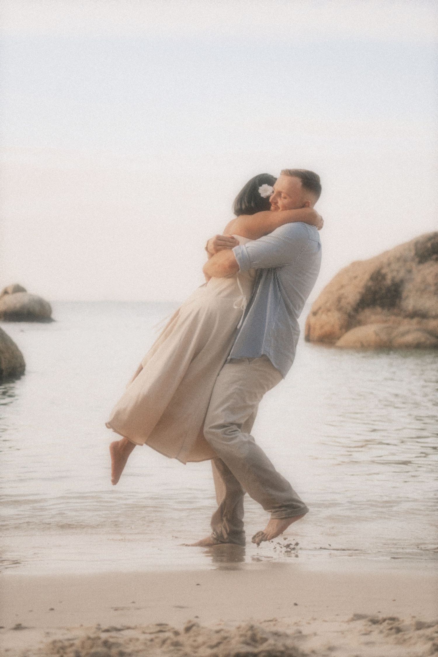 Man lifting a woman in a flowing dress as they laugh in the shallows