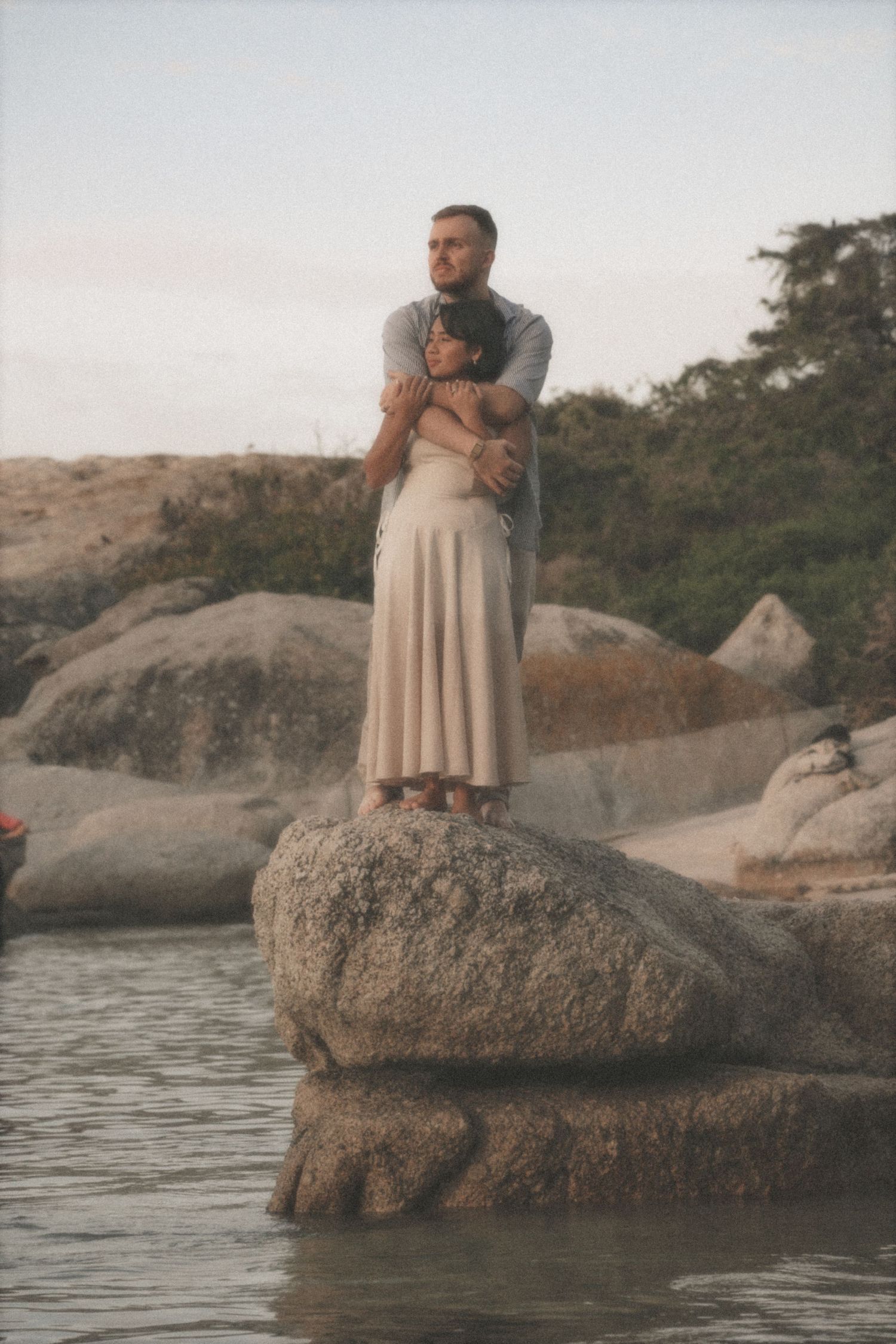 Couple on a large boulder in the sea as he wraps his arms around her, Cape Town couple's session