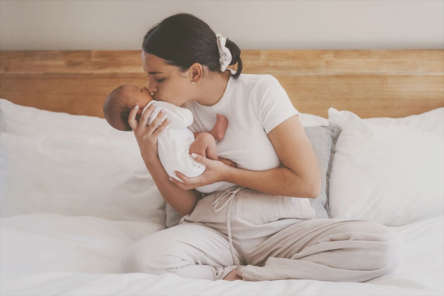 Mother sitting cross-legged on a bed kissing her newborn, Cape Town lifestyle session at home