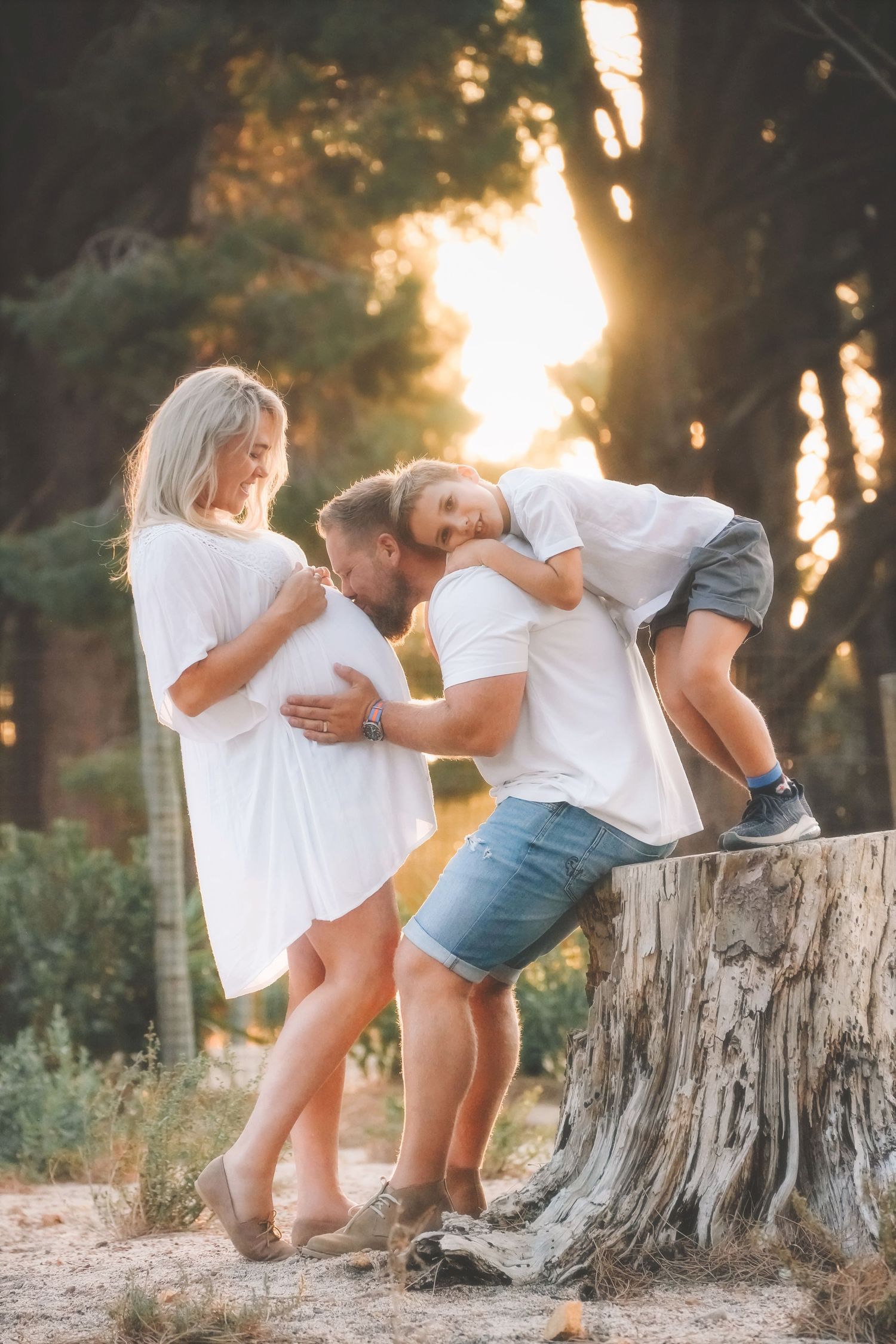 Parents-to-be and their young son laughing beside a tree stump in golden light