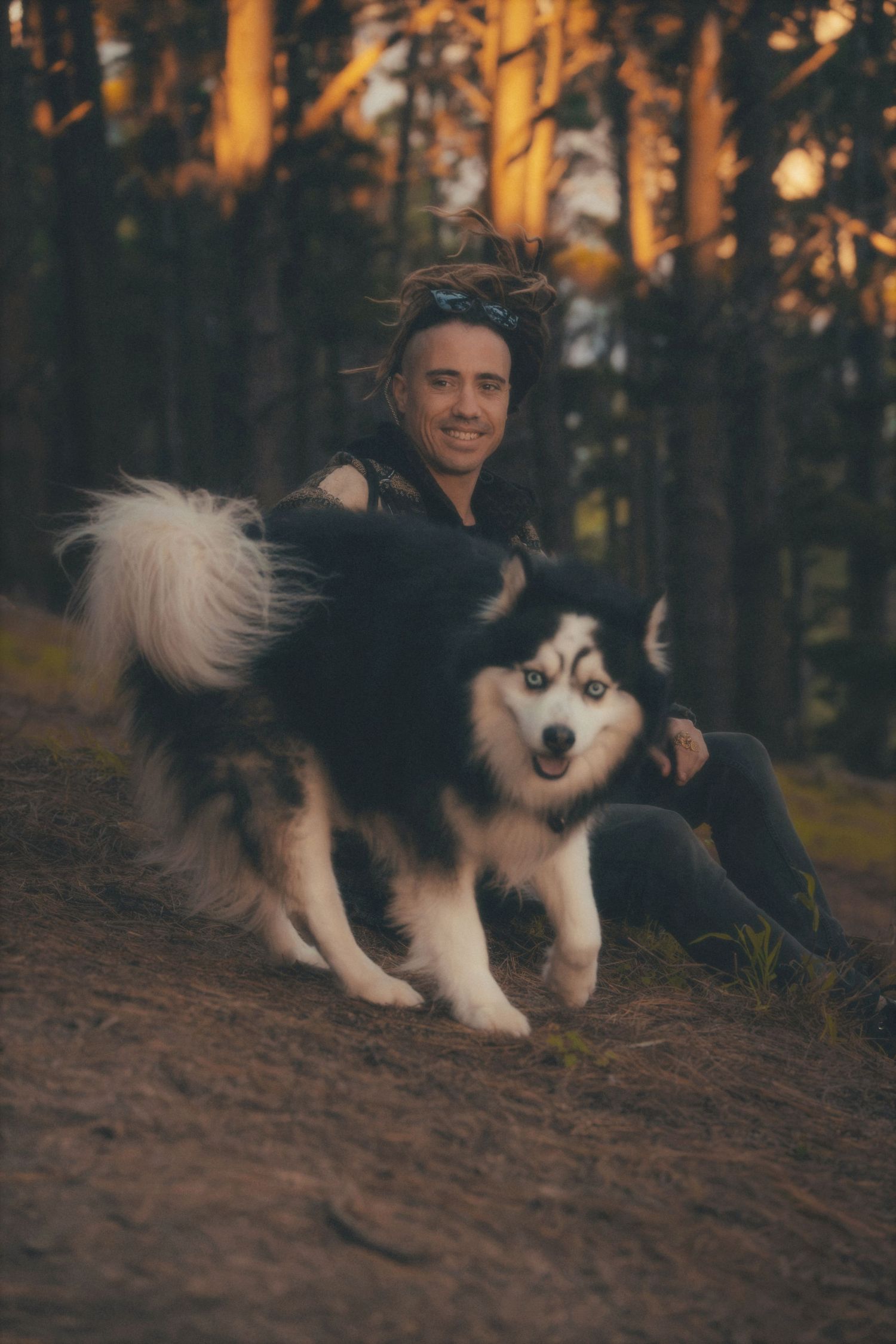 Man sitting in the pines with his husky in front of him