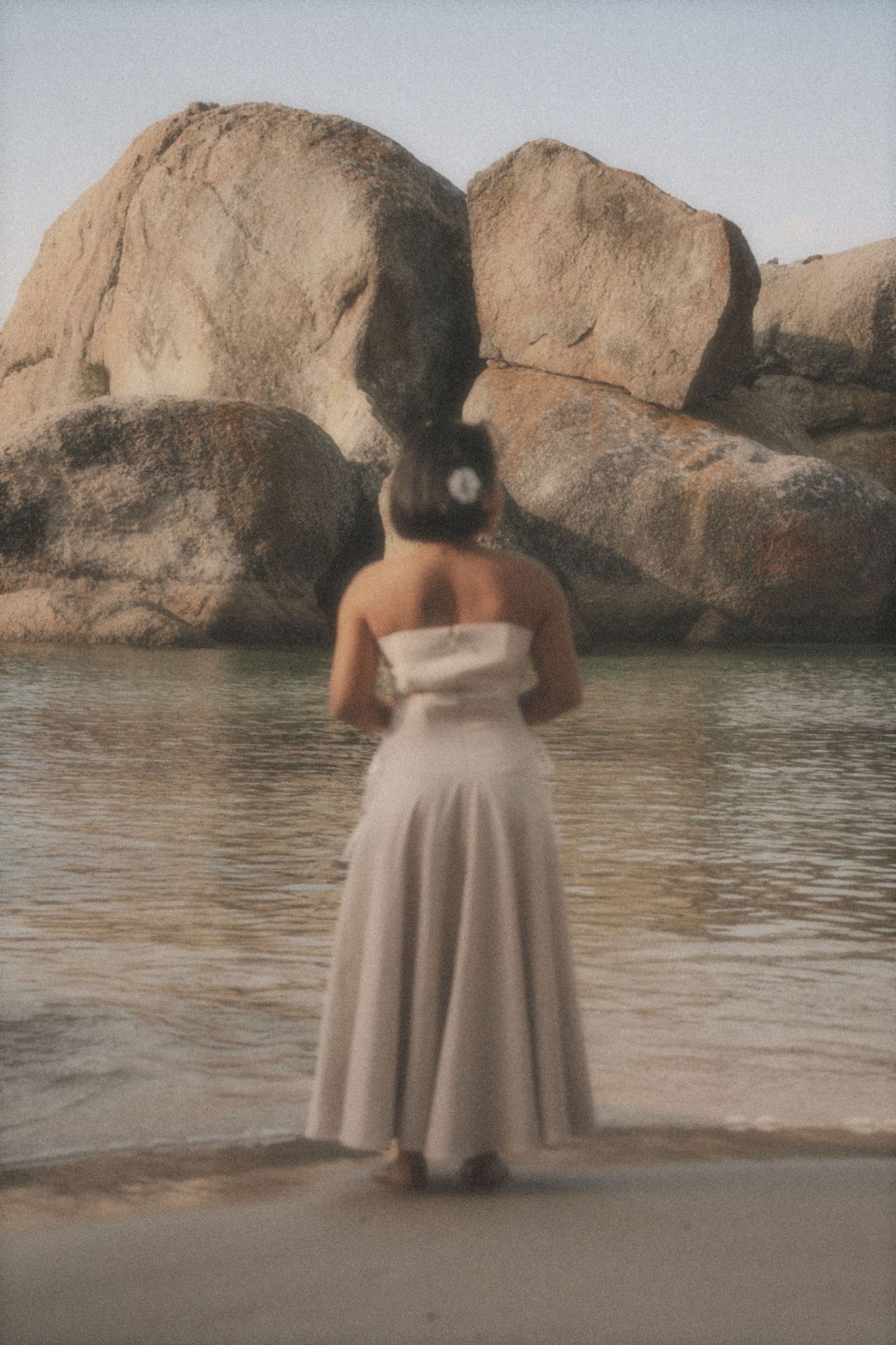 Woman in a cream dress with a flower in her hair, seen from behind, facing the sea among large boulders