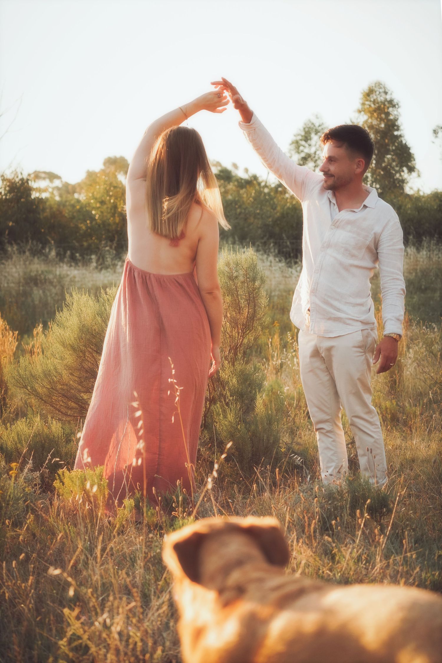 Couple dancing in a field at golden hour as she twirls under his arm, a dog in the foreground