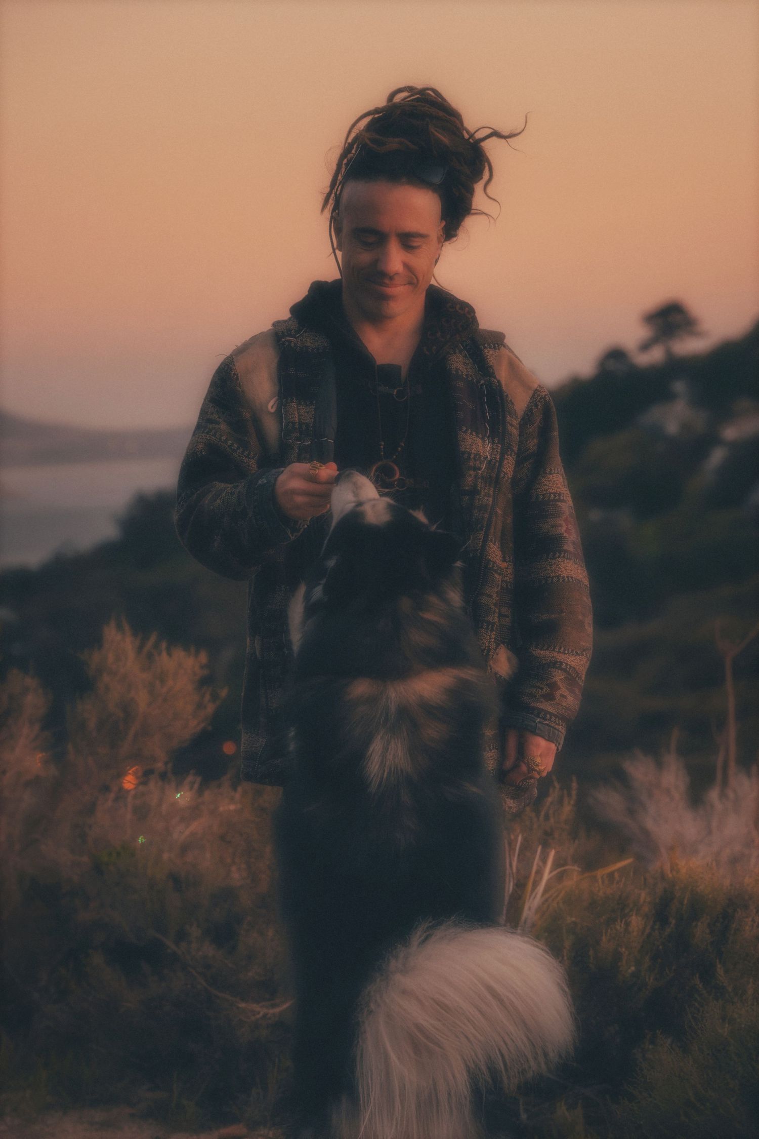 Man smiling down at his dog against a soft sunset sky and the sea