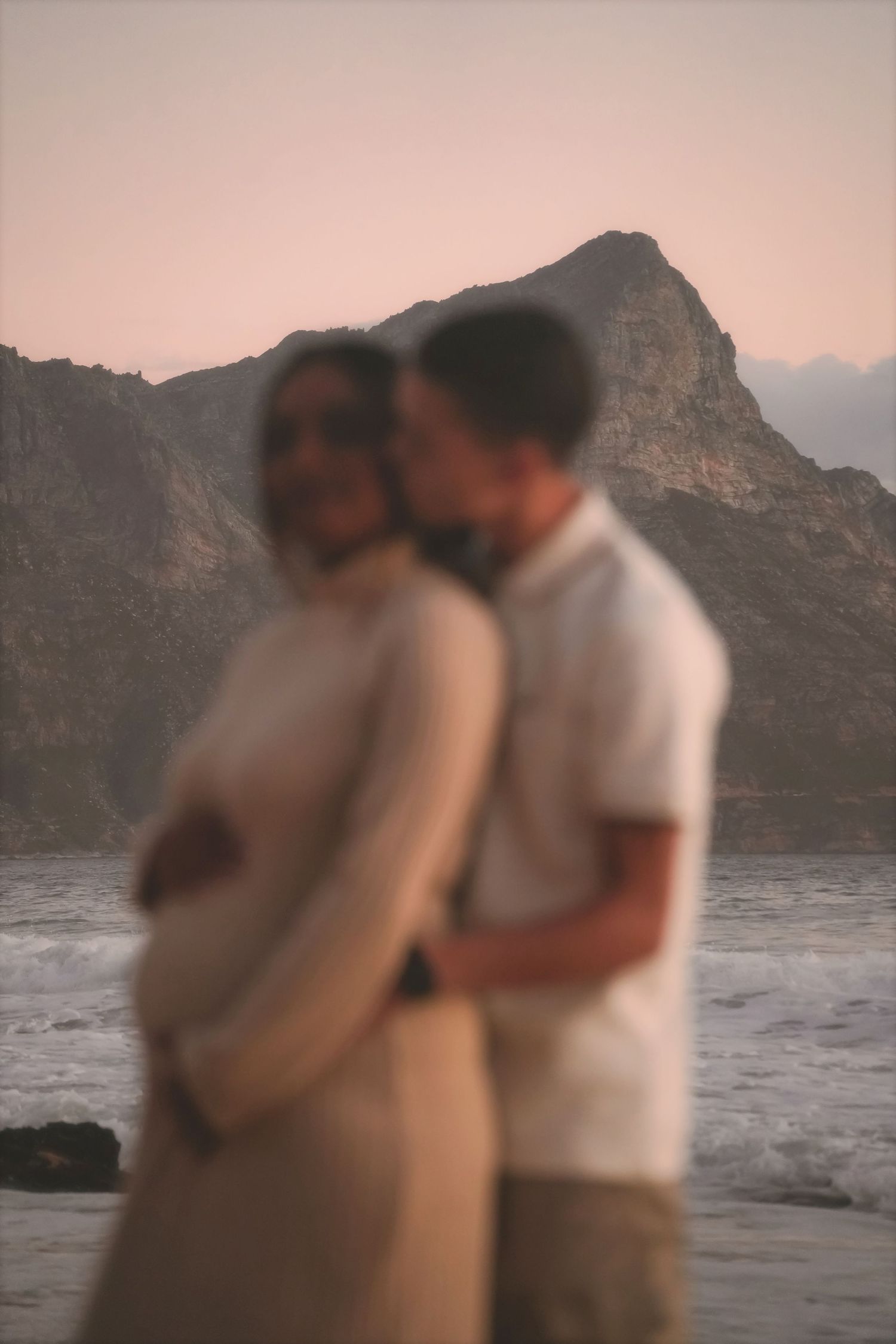 Expecting couple embracing in soft focus, hands on her bump, mountains and sea behind