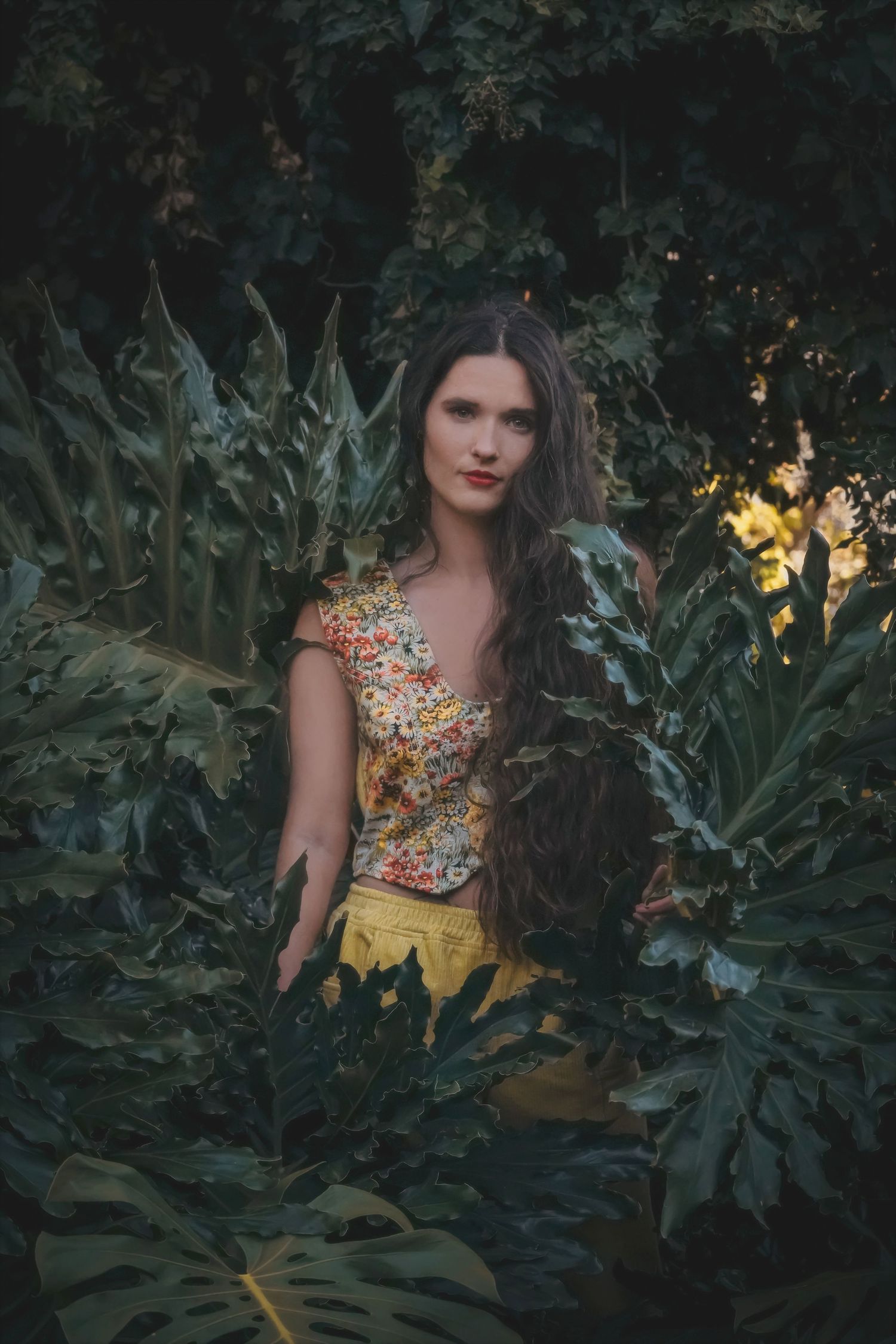 Woman in a floral vest and yellow trousers standing among large monstera leaves