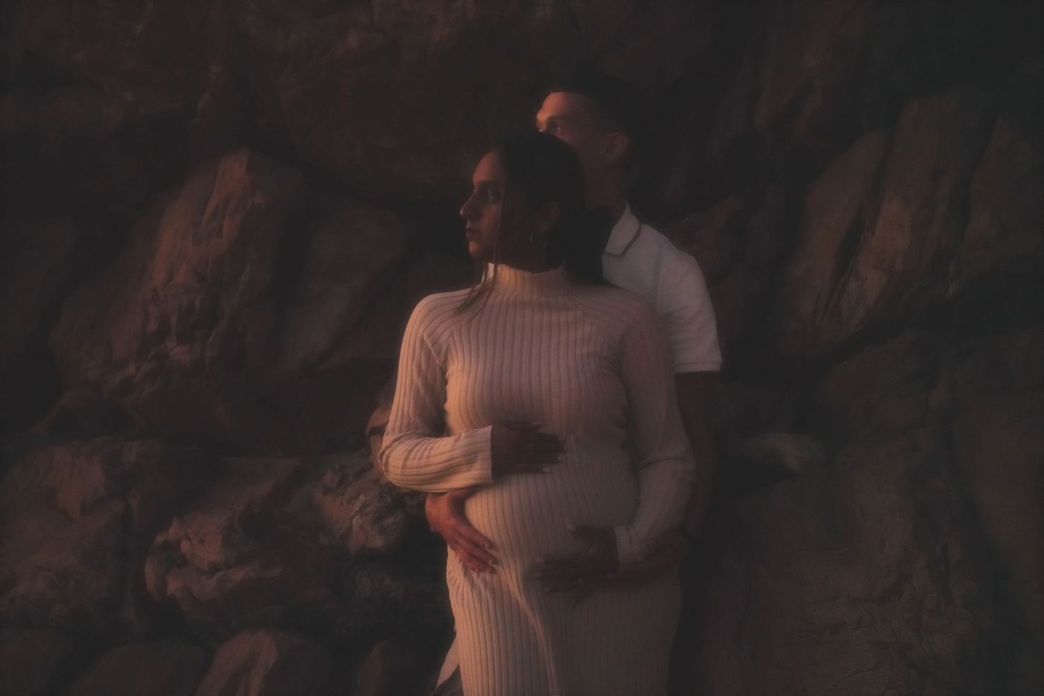 Couple standing close against a dark rock wall, her hand resting on her baby bump