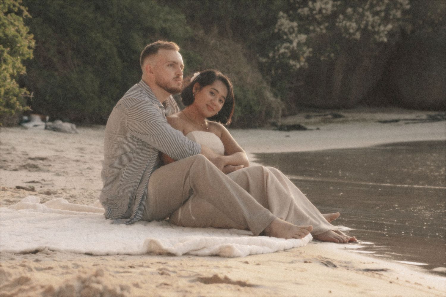 Couple sitting together on a blanket on the sand beside coastal trees