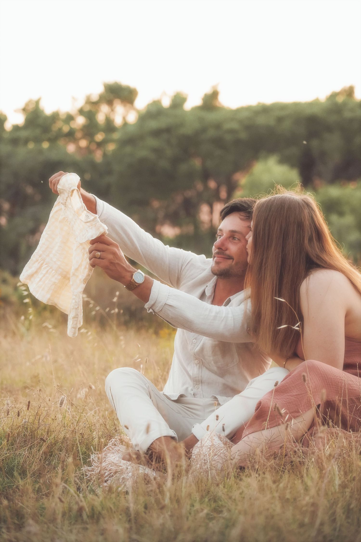 Expecting couple sitting in the grass, holding up a tiny baby outfit
