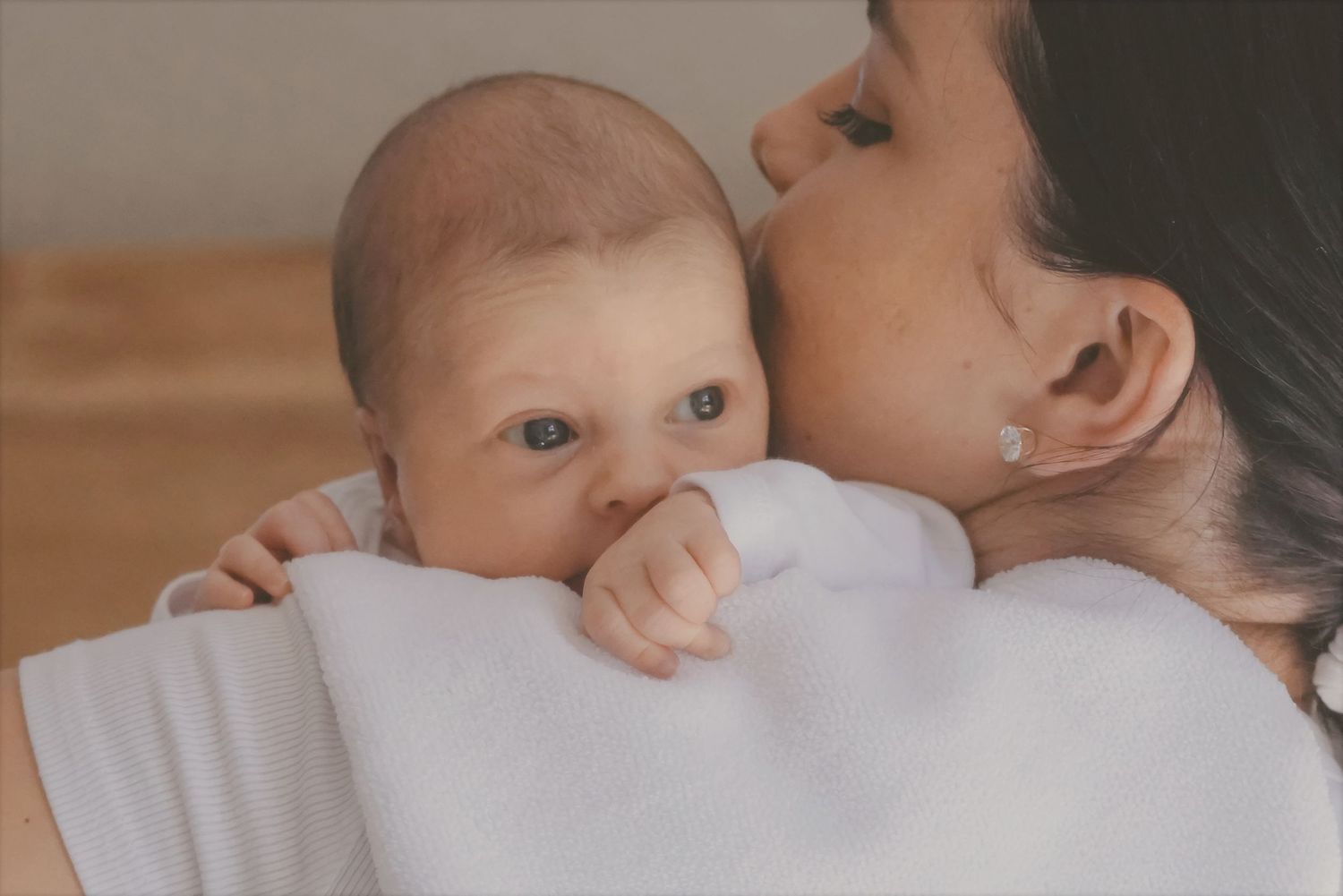Newborn peeking over mum's shoulder as she kisses their cheek, a wooden headboard behind