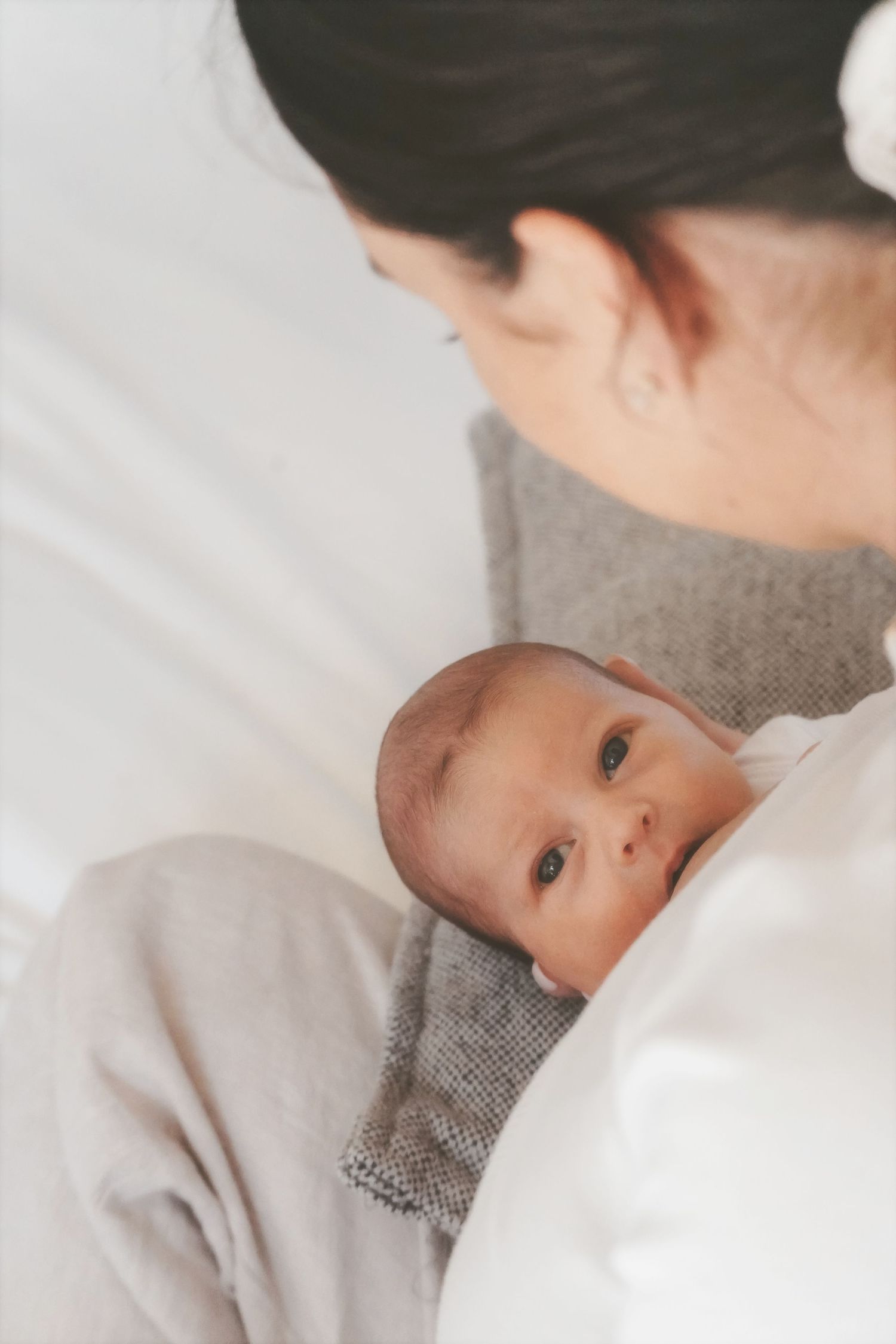 Newborn looking up from mum's chest on a white bed in soft natural light
