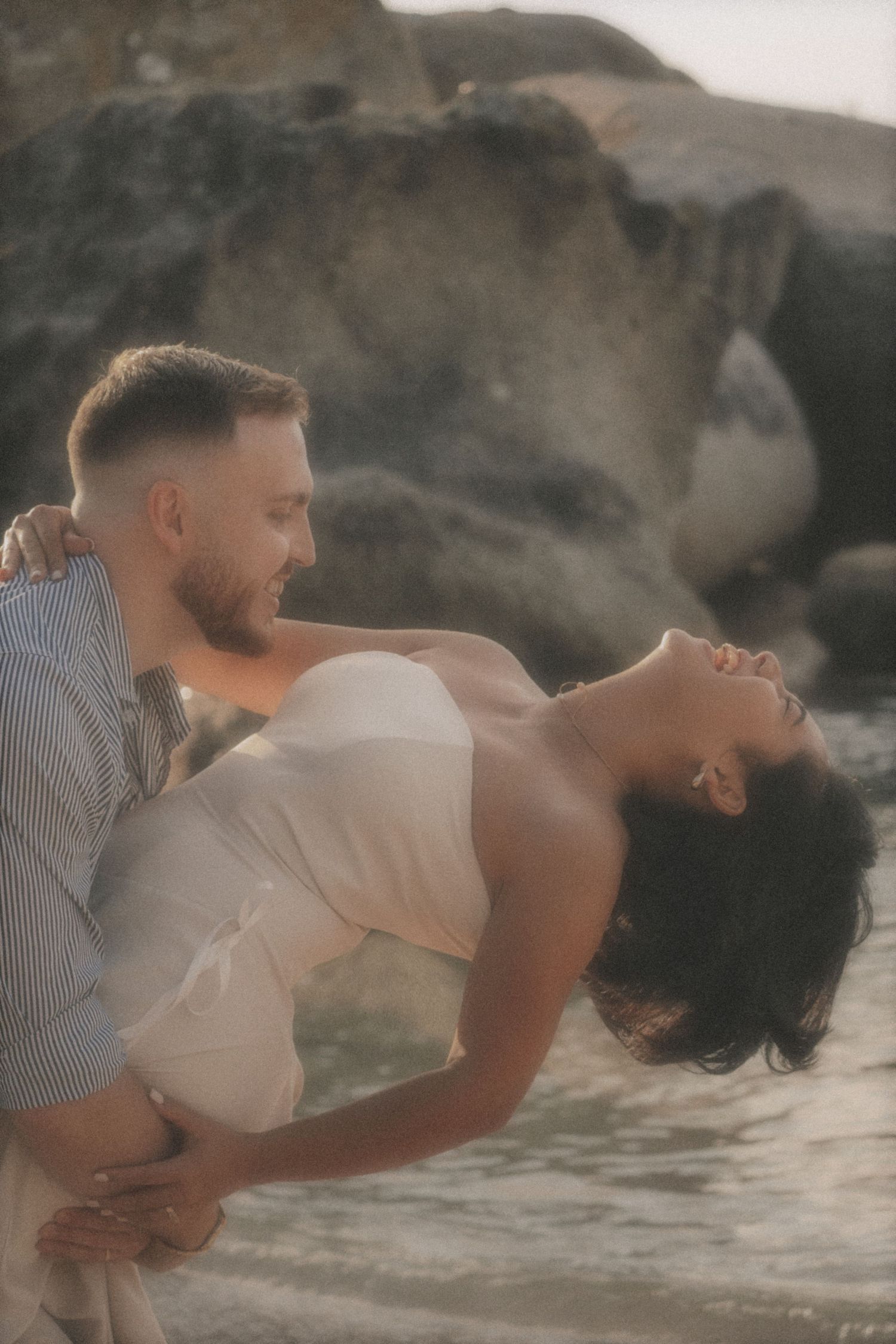 Man dipping a woman backwards as she laughs, in shallow water among rocks