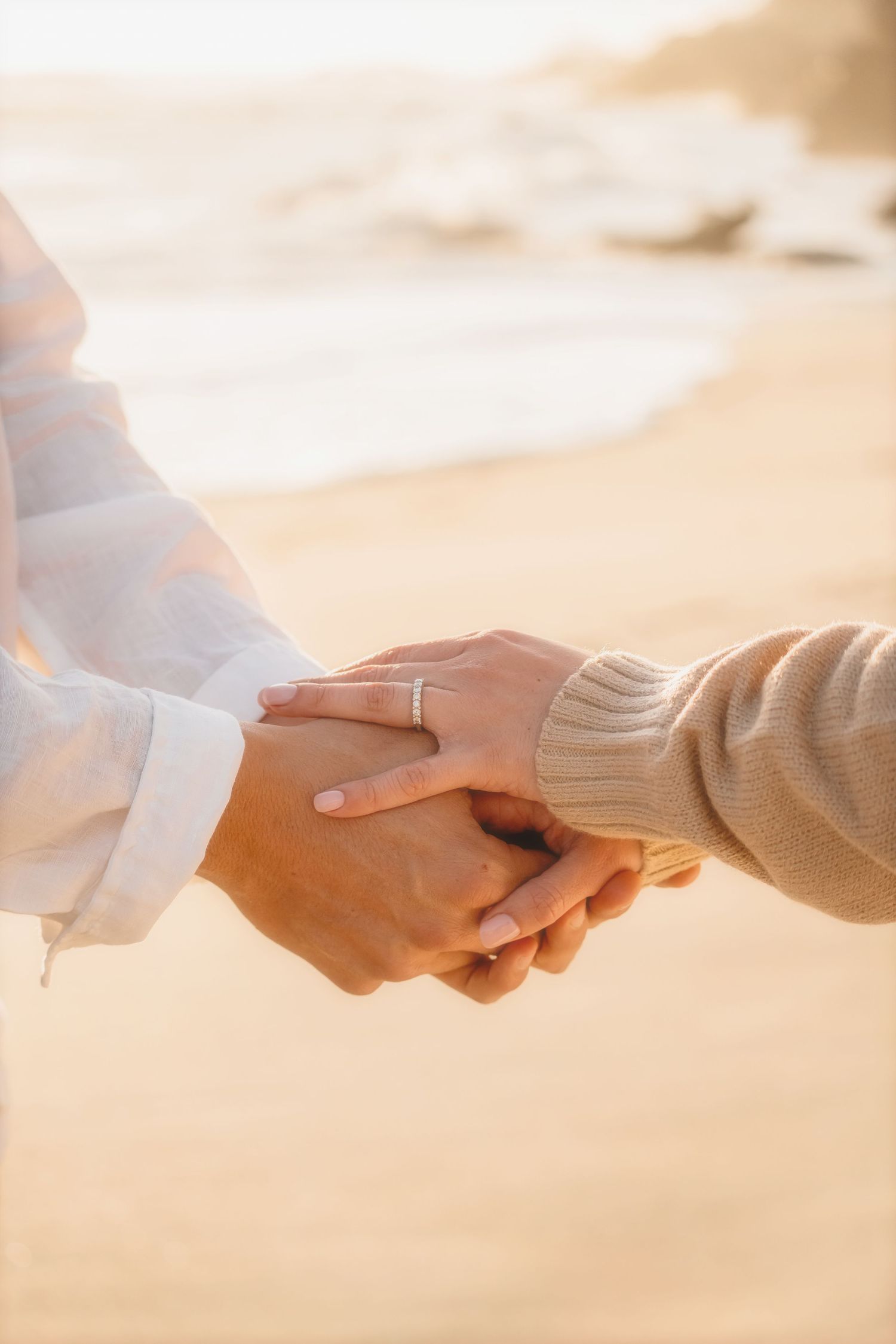 Close-up of clasped hands, an engagement ring on her finger, on a golden beach