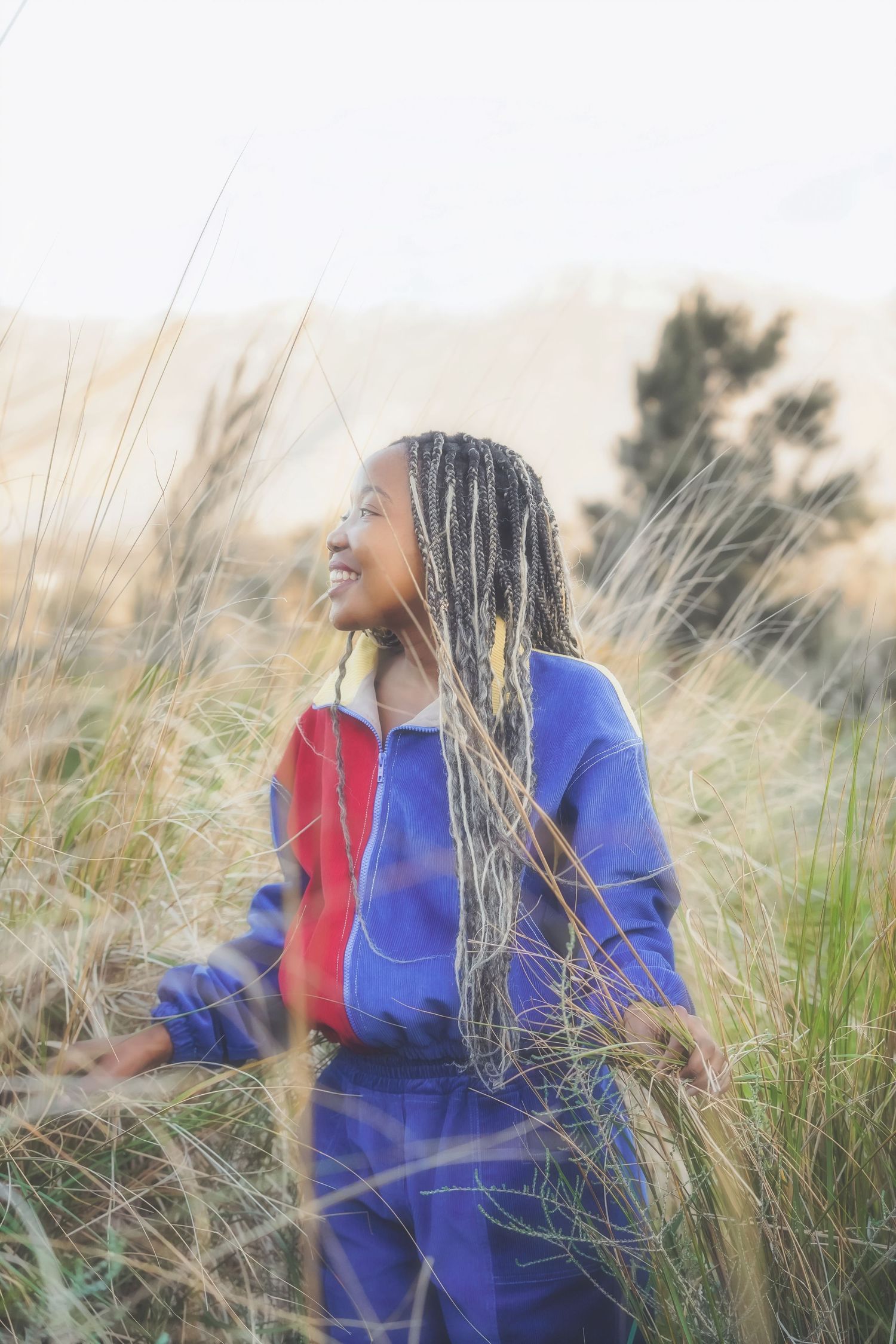 Woman with long braids in a blue and red jacket smiling up in tall grass