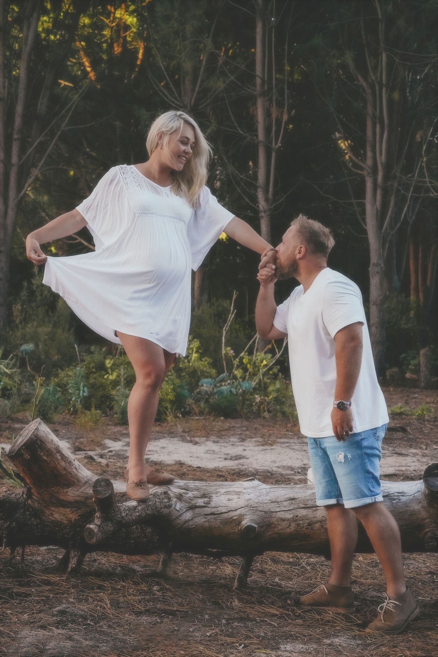 Pregnant woman lifting her white dress as her partner kisses her hand beside a fallen log