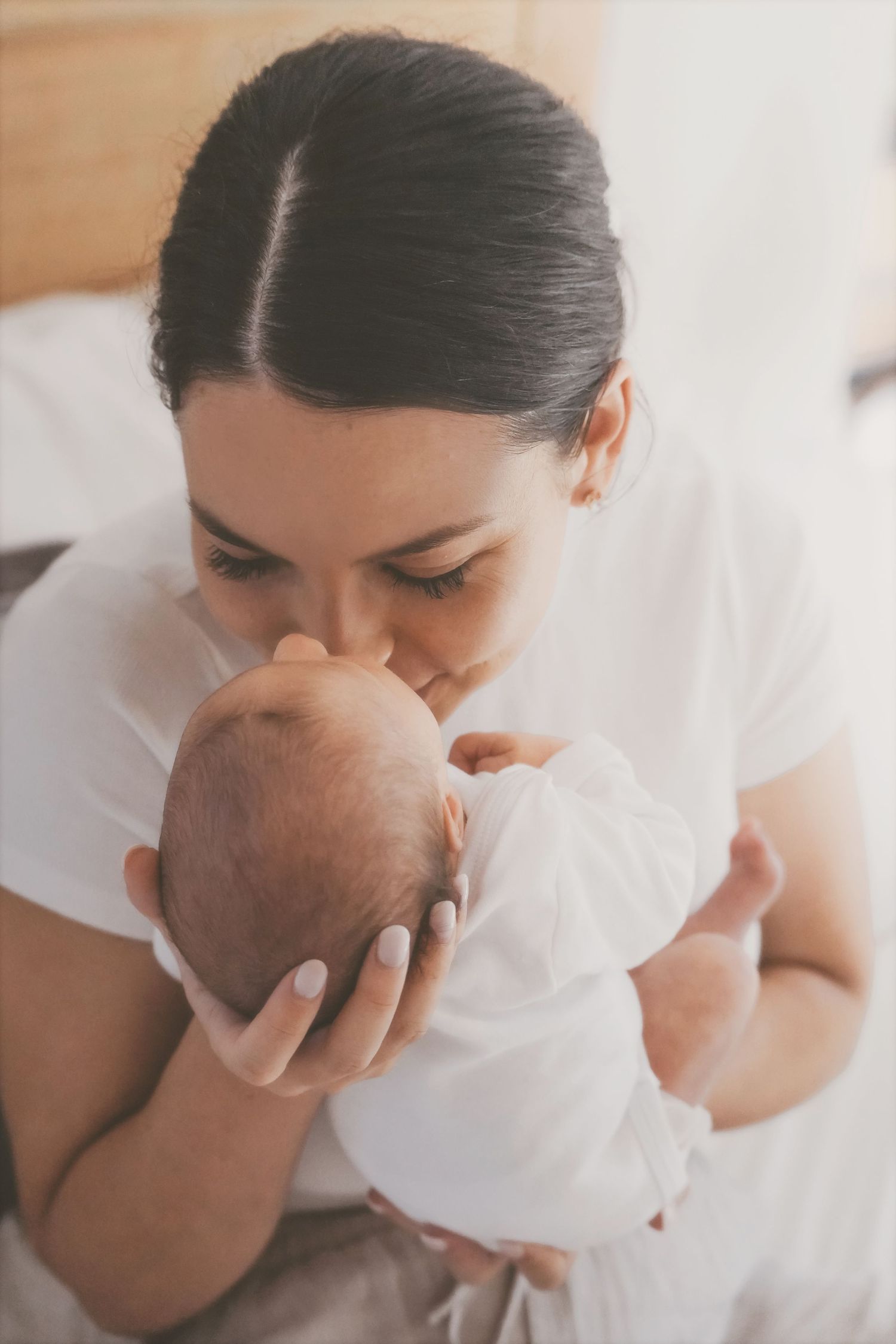Mother kissing the top of her newborn's head as she holds them close in soft window light