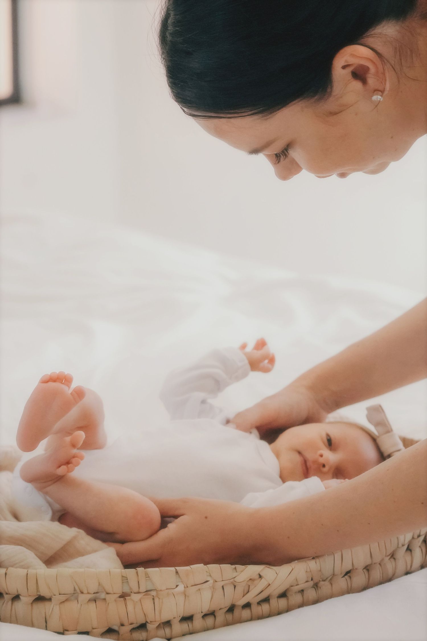 Wide shot of a swaddled newborn in a bow headband on a white bed beside a crocheted toy