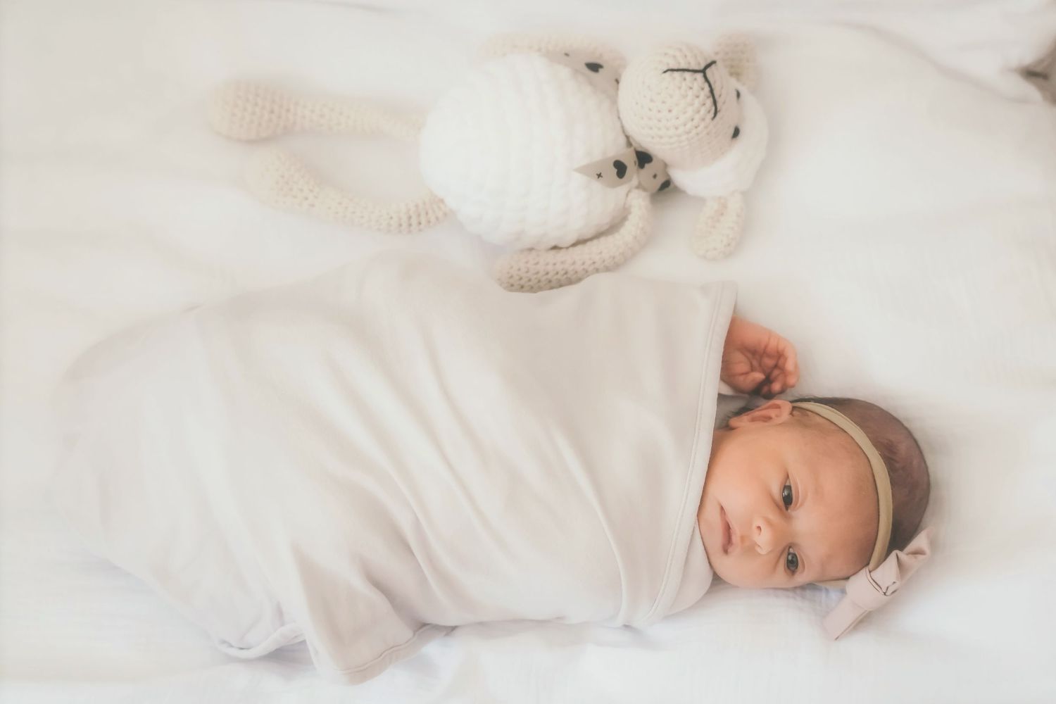 Newborn lying on a white bed with tiny hands up to their mouth, looking at the camera