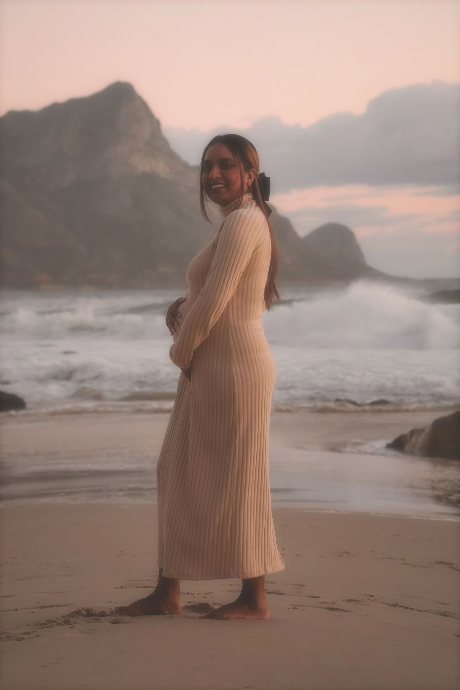 Smiling pregnant woman in a ribbed cream knit dress standing barefoot on the shore with surf and mountains behind
