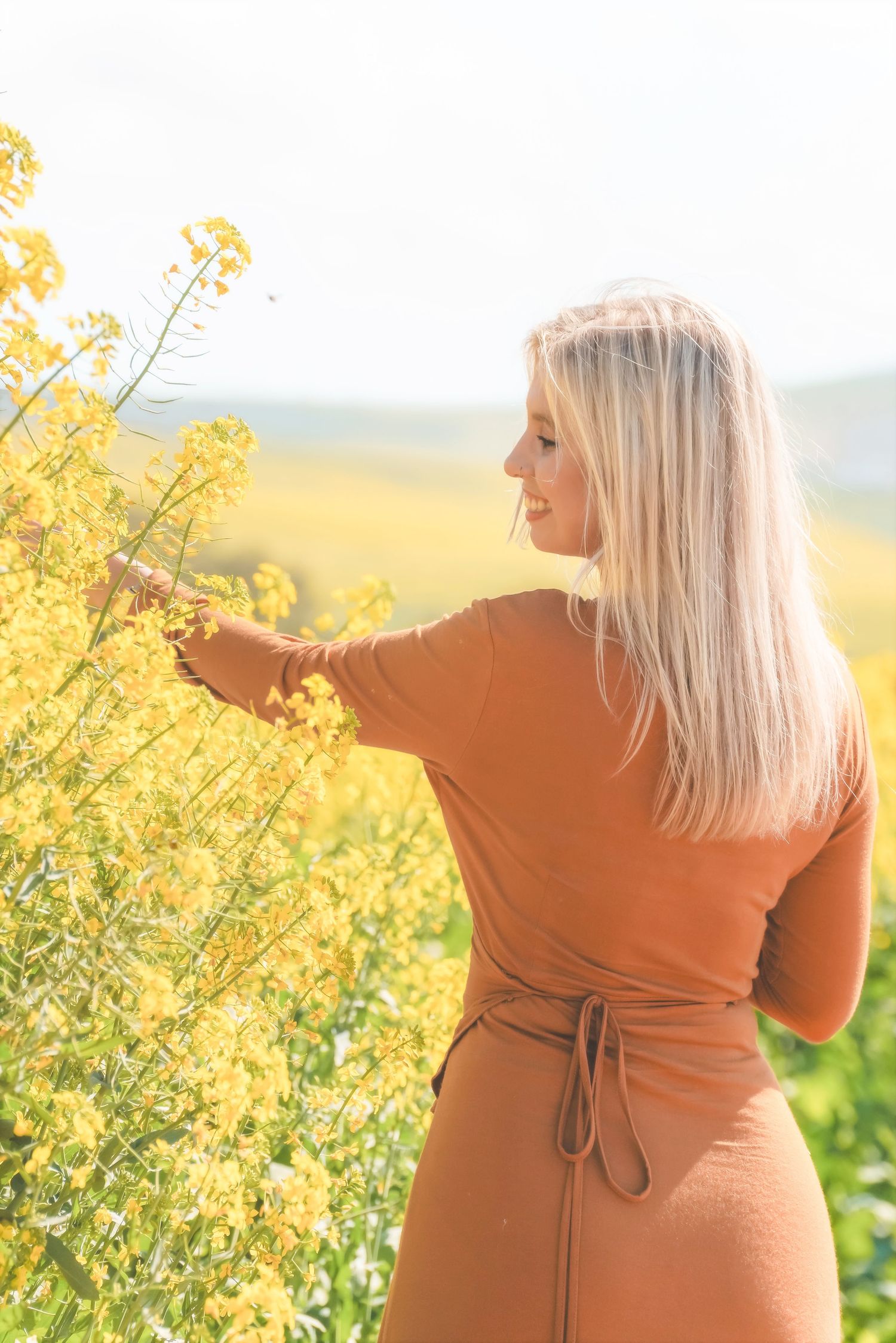 Smiling woman in a rust dress reaching into the canola in profile