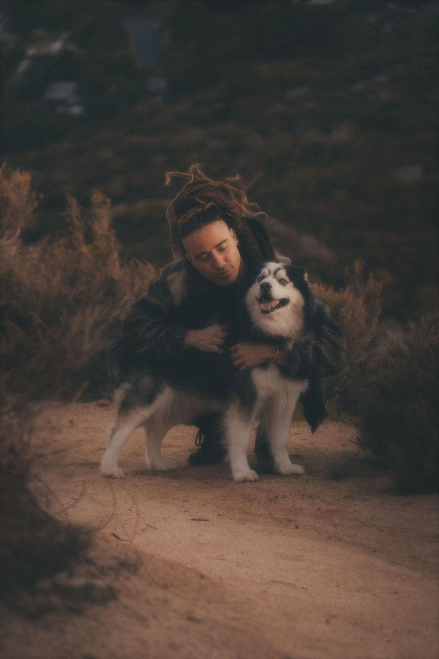 Man crouching to hug his husky on a sandy trail with fynbos behind them