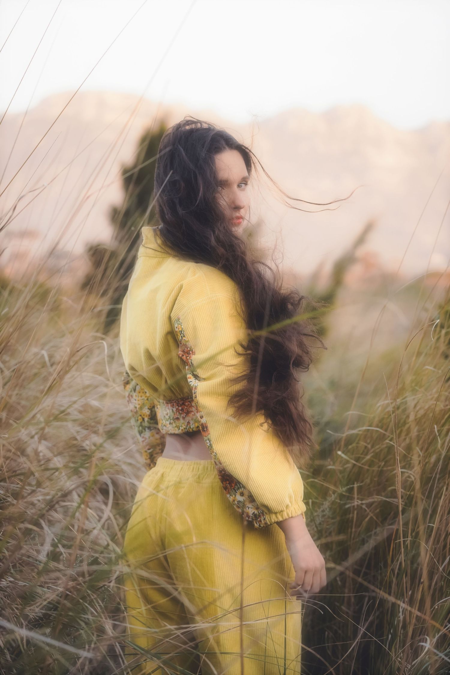 Woman in a yellow jacket looking back, hair blowing, with mountains behind