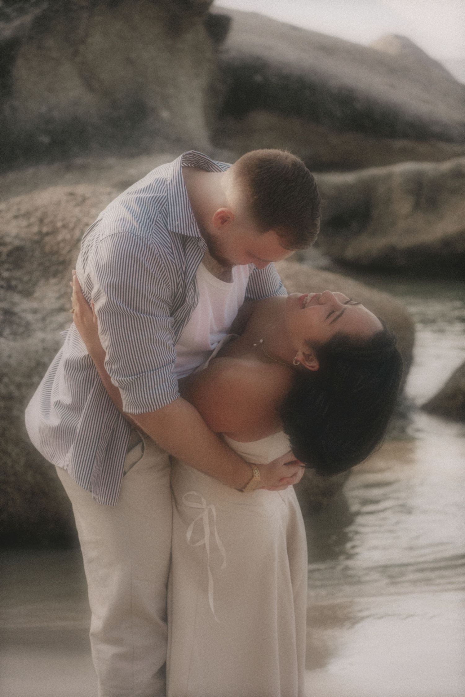 Man in a striped shirt dipping a woman in a cream dress in shallow water beside boulders