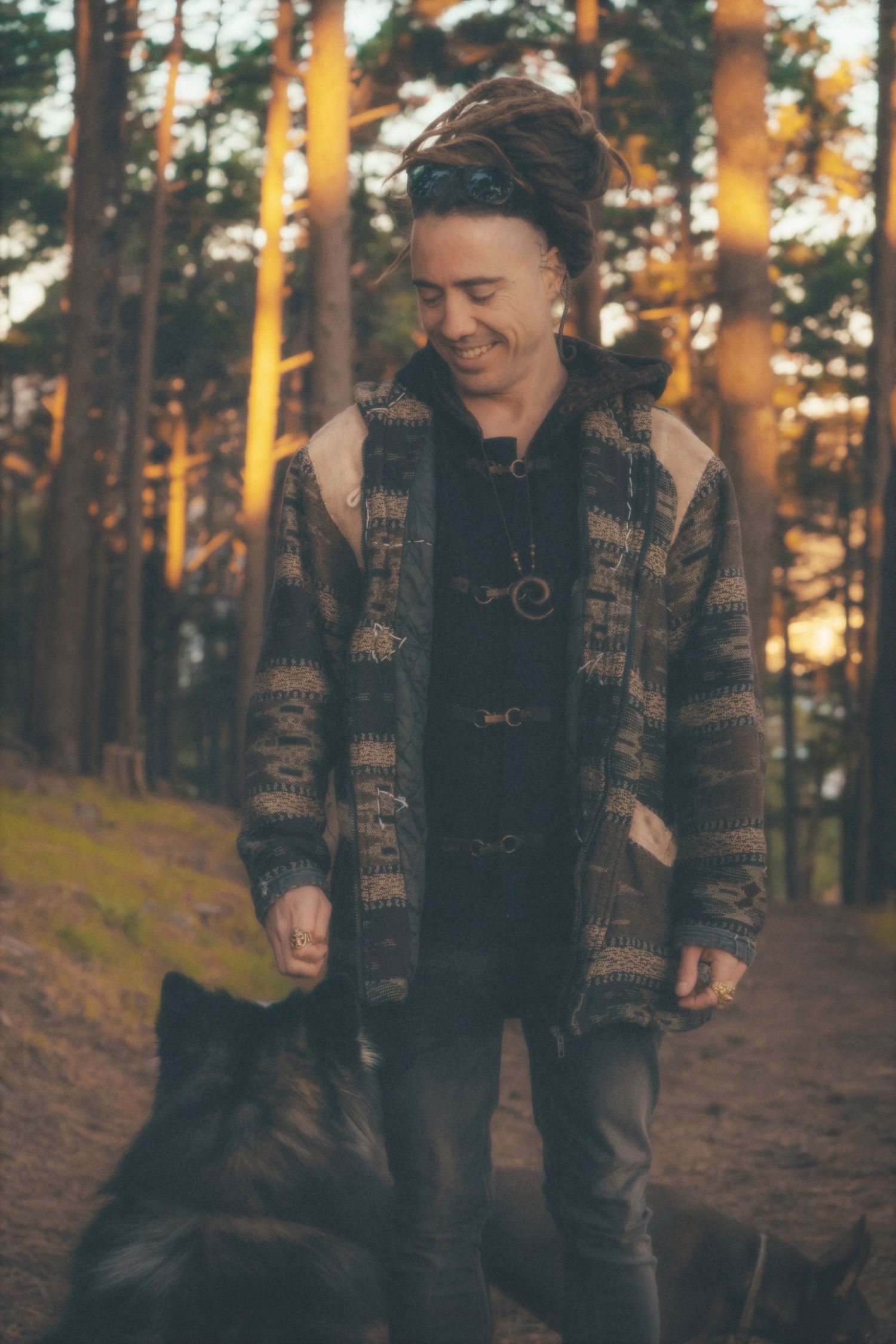 Smiling man in a patterned jacket looking down at his dog among pine trees