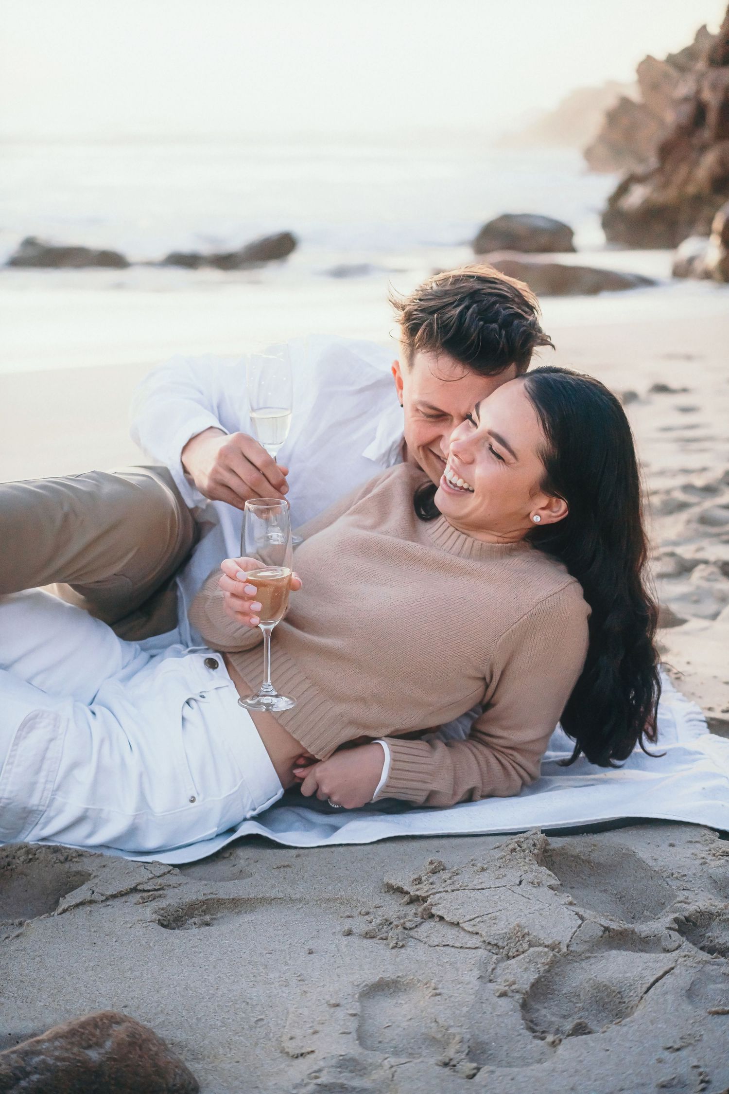 Couple laughing on a picnic blanket on the sand, holding champagne glasses