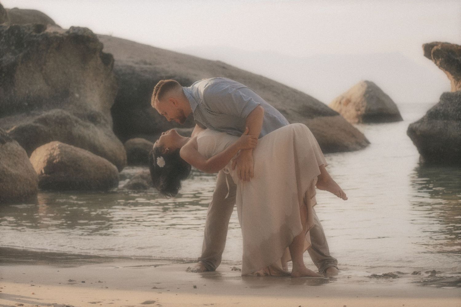 Wide shot of a couple in shallow water among rocks as he bends over her