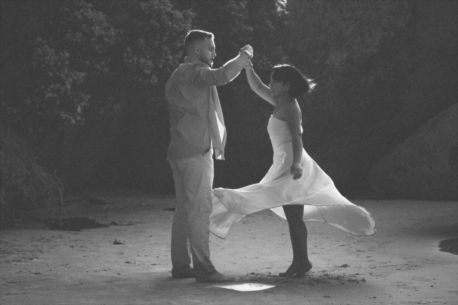 Black and white photo of a couple dancing on the sand as her dress swirls