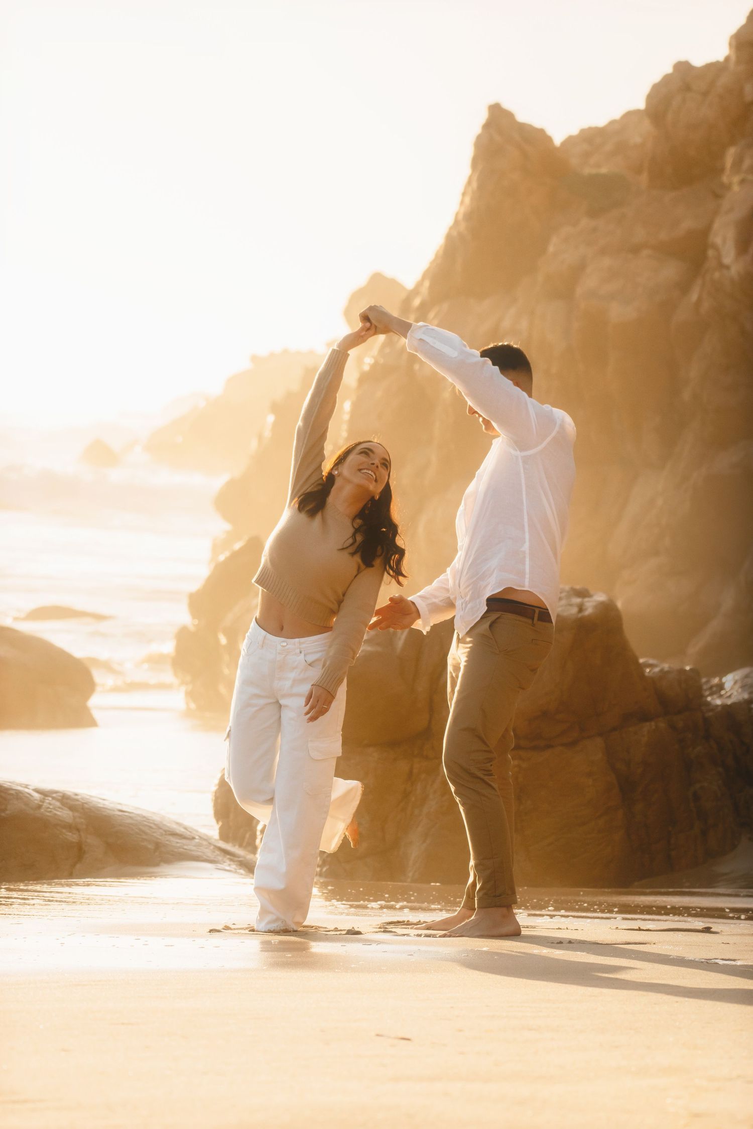 Woman twirling under his arm as the couple dance on the sand beside sunlit rocks