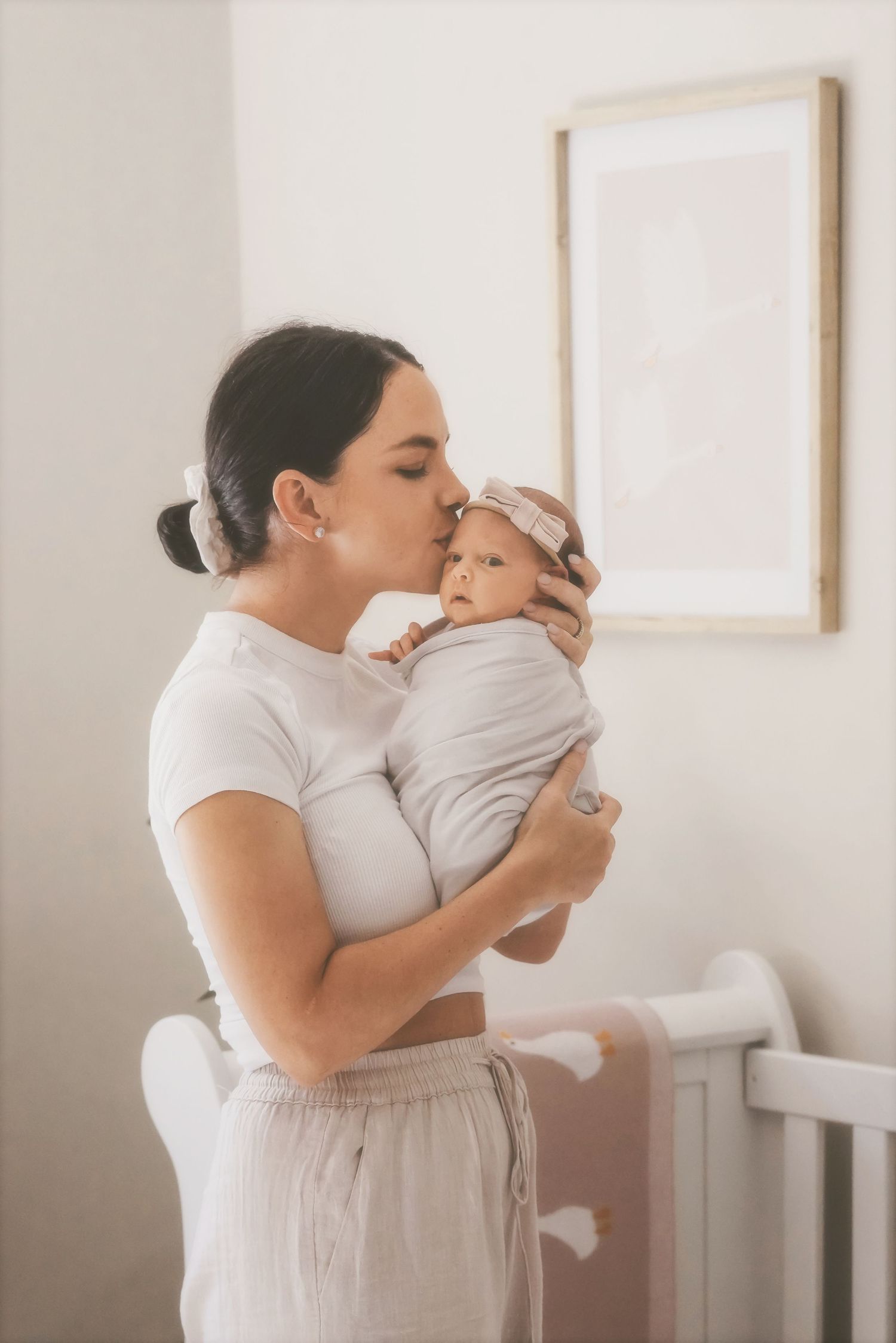Mother kissing her swaddled newborn beside a white crib in a soft, neutral nursery