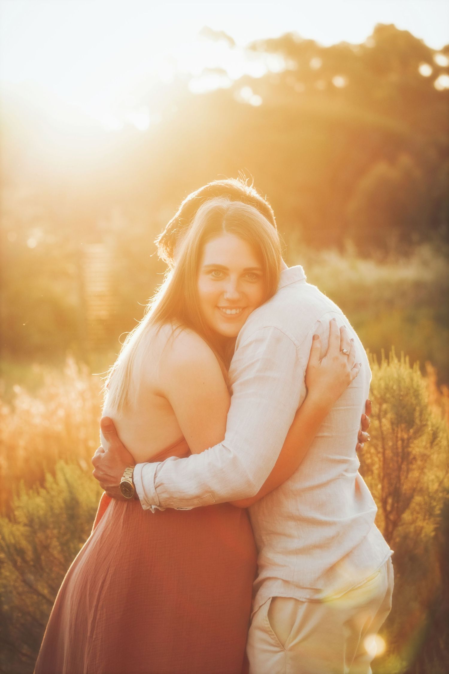 Smiling woman in a terracotta dress hugging her partner in backlit golden light