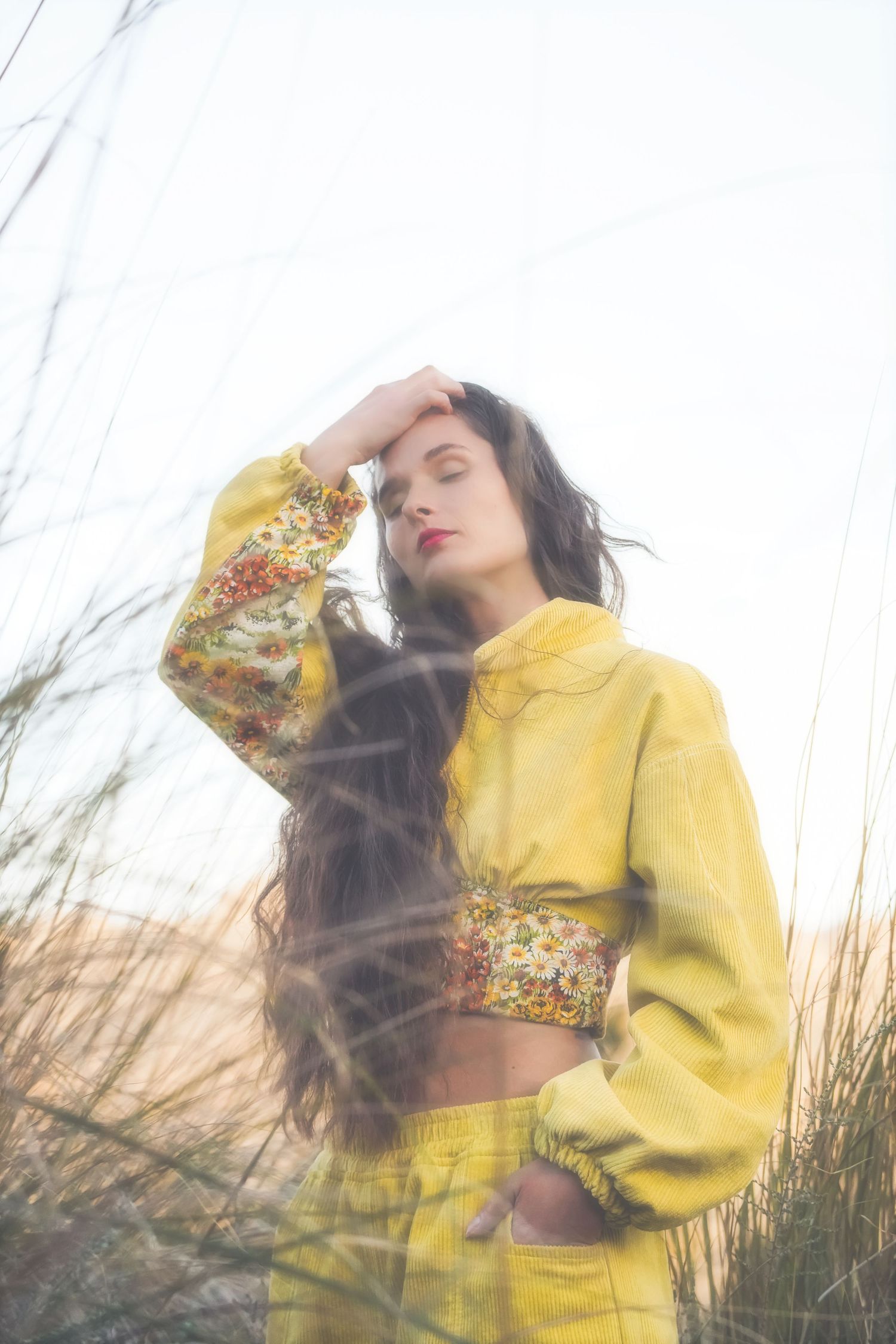 Woman in yellow with eyes closed and a hand in her hair, tall grass around her