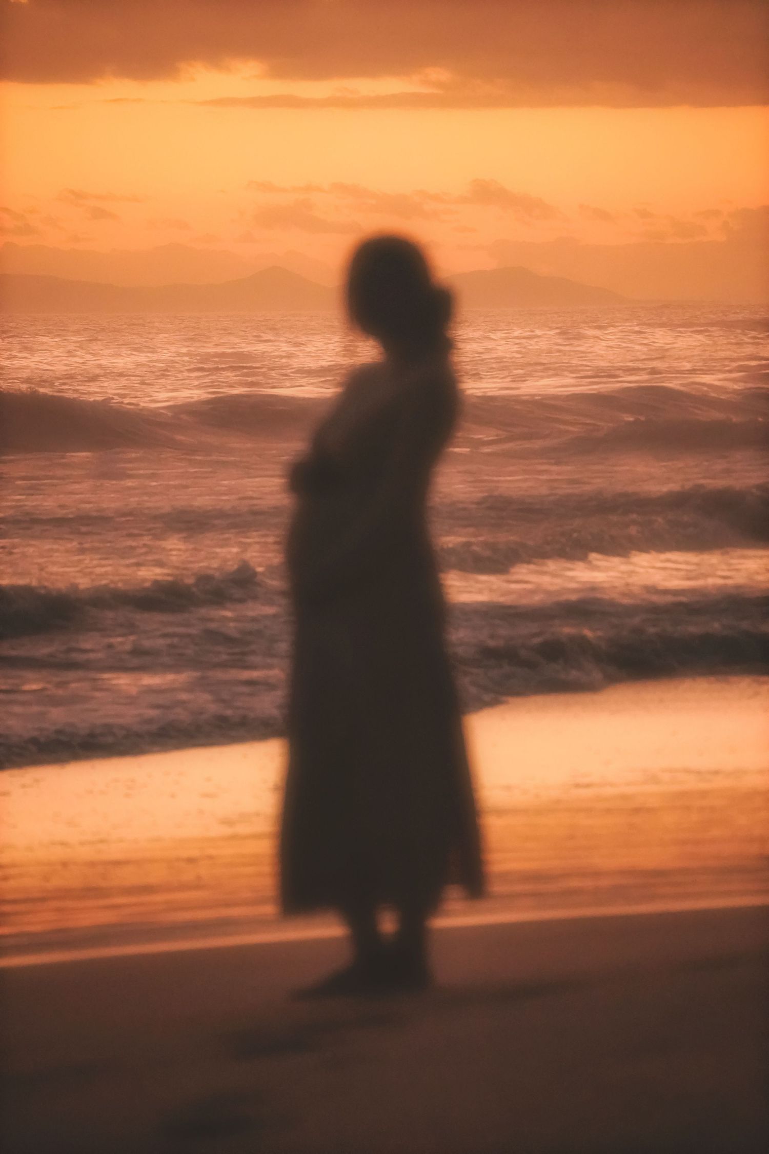 Soft, blurred silhouette of a pregnant woman in the surf under a glowing orange sky