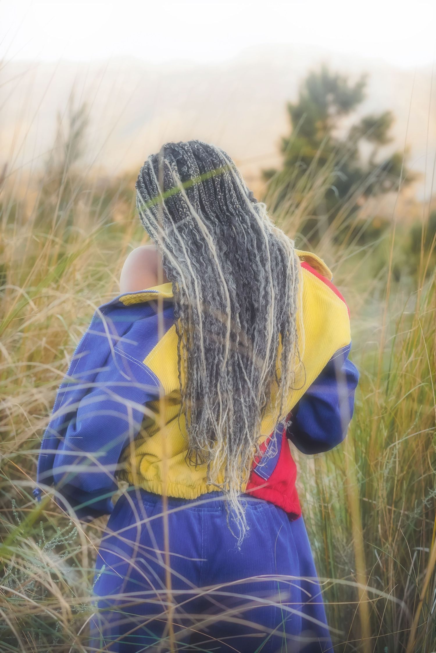 Woman with long braids seen from behind in a blue and yellow jacket among tall grass