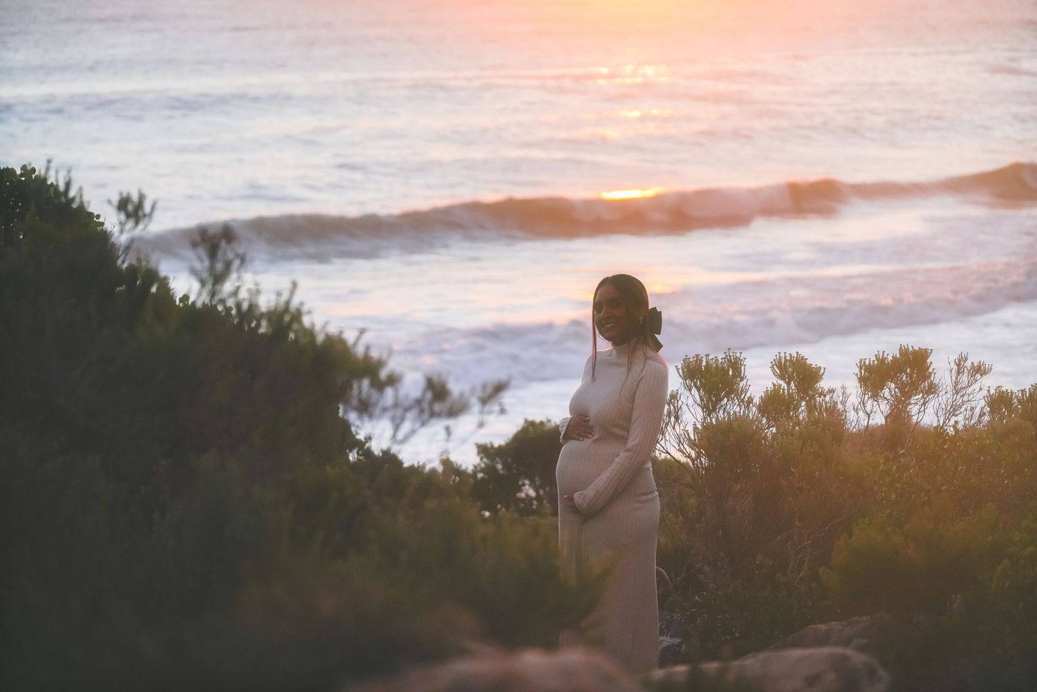 Woman in a long cream dress standing among coastal bushes looking out over the sea at sunset