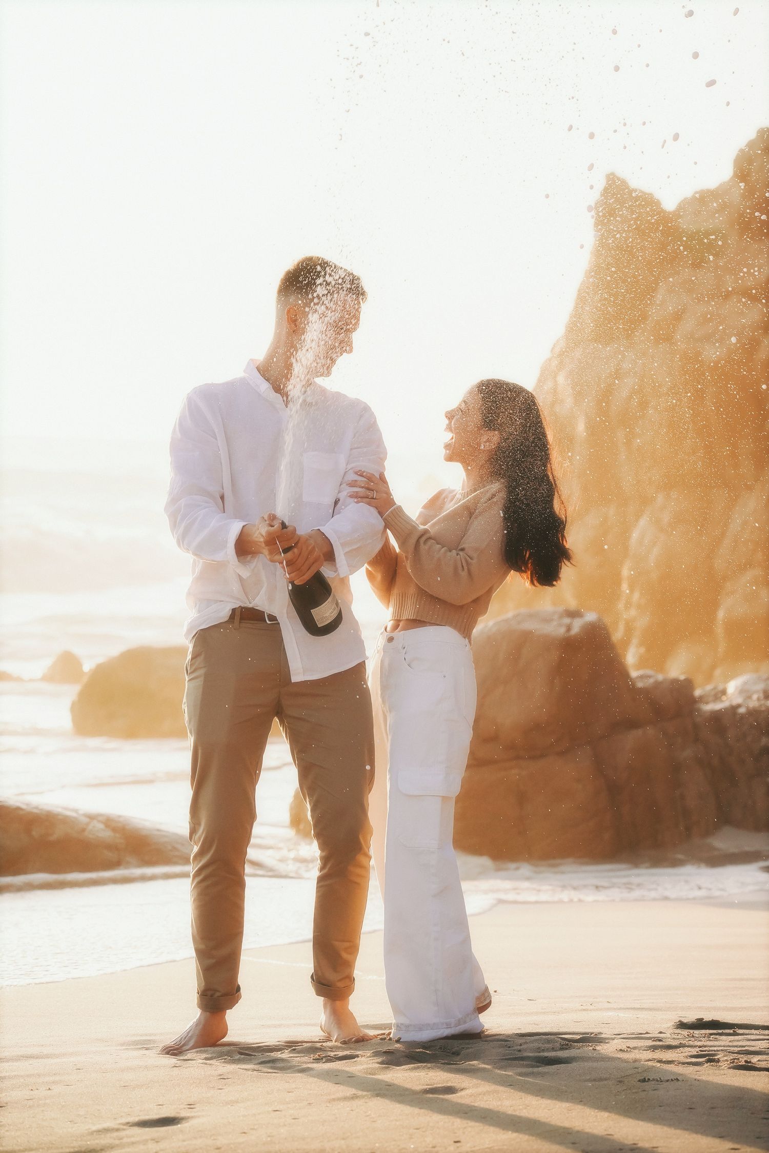 Couple on the beach at golden hour as he pops a champagne bottle, spray catching the light
