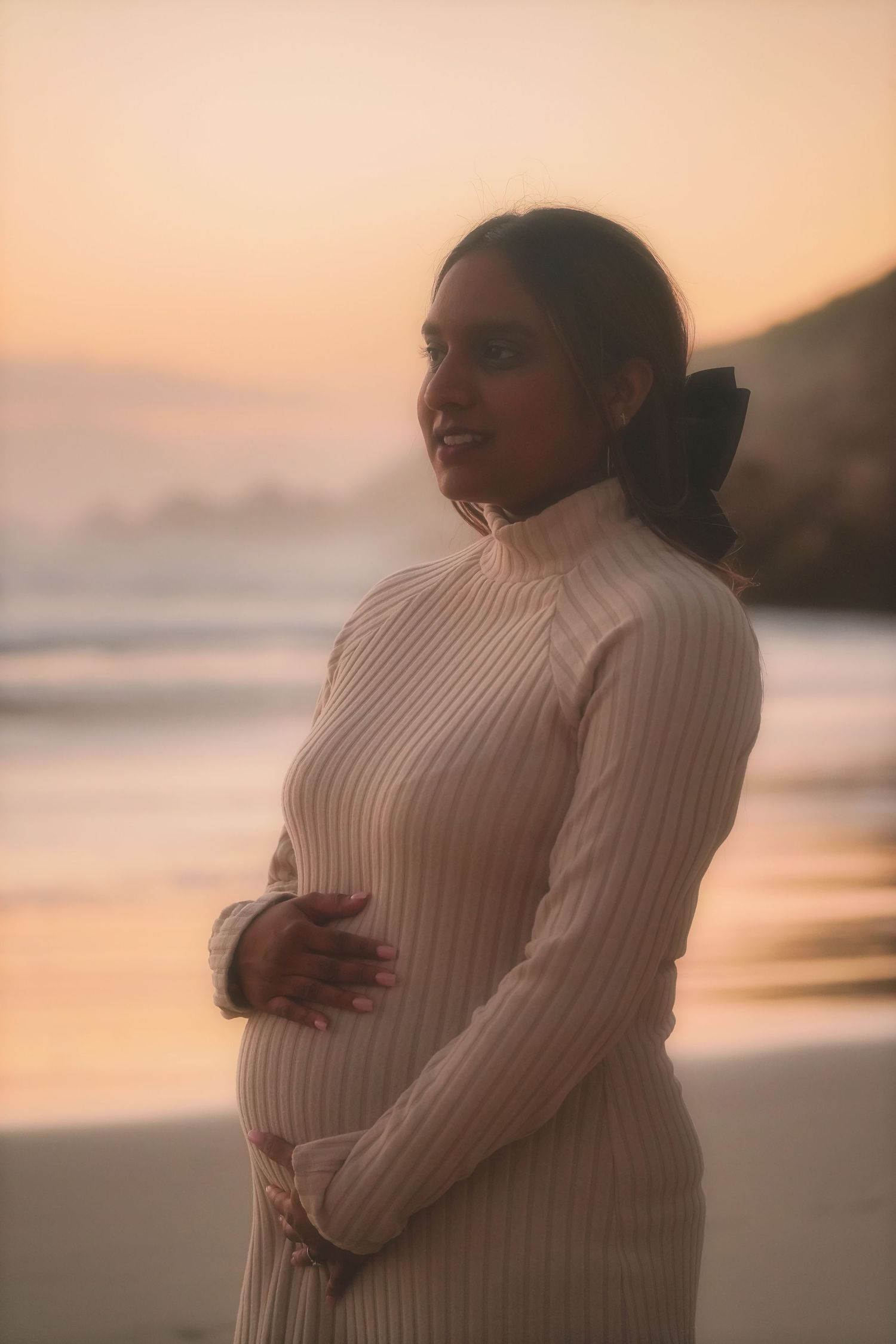 Pregnant woman in a cream knit with a bow in her hair, hands on her bump, gazing out at a Cape Town sunset