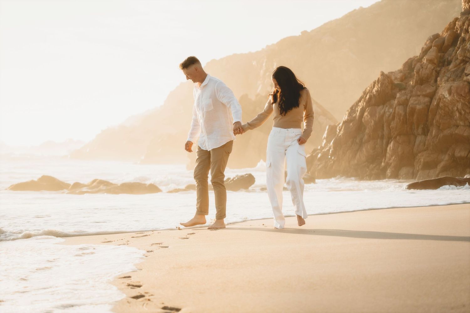 Couple walking hand in hand along a Cape Town shoreline below a rocky headland