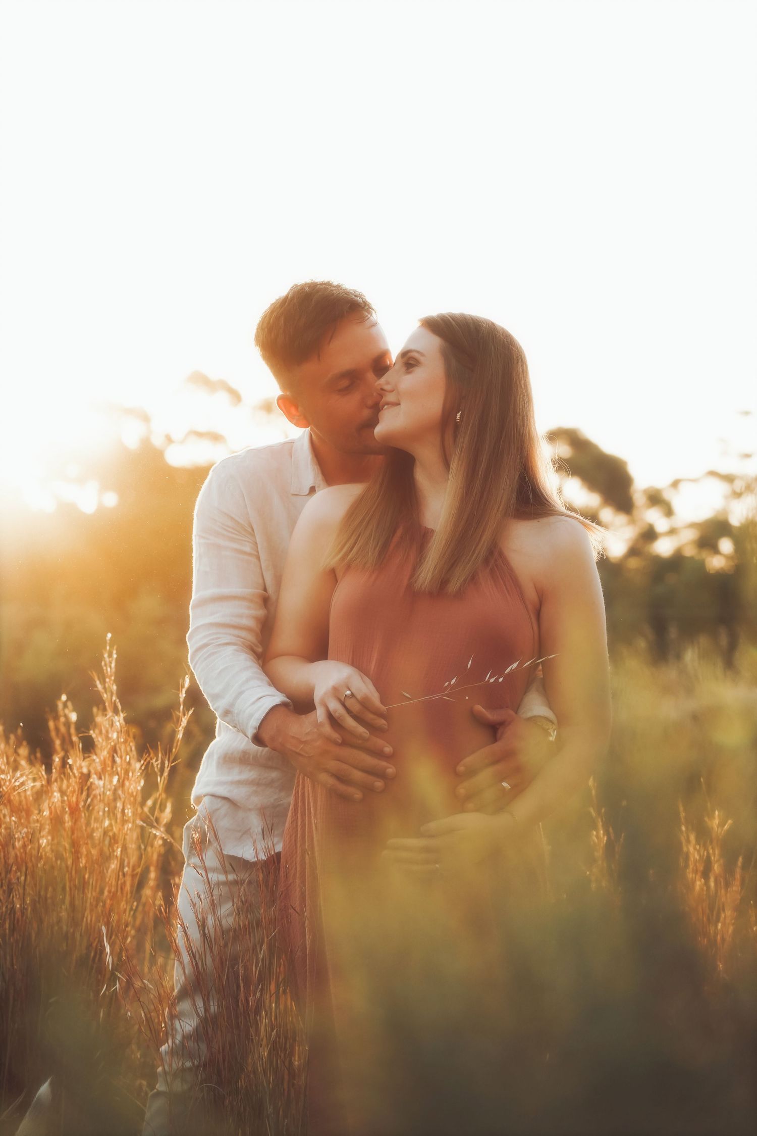 Couple kissing in tall golden grass, his hands resting on her baby bump