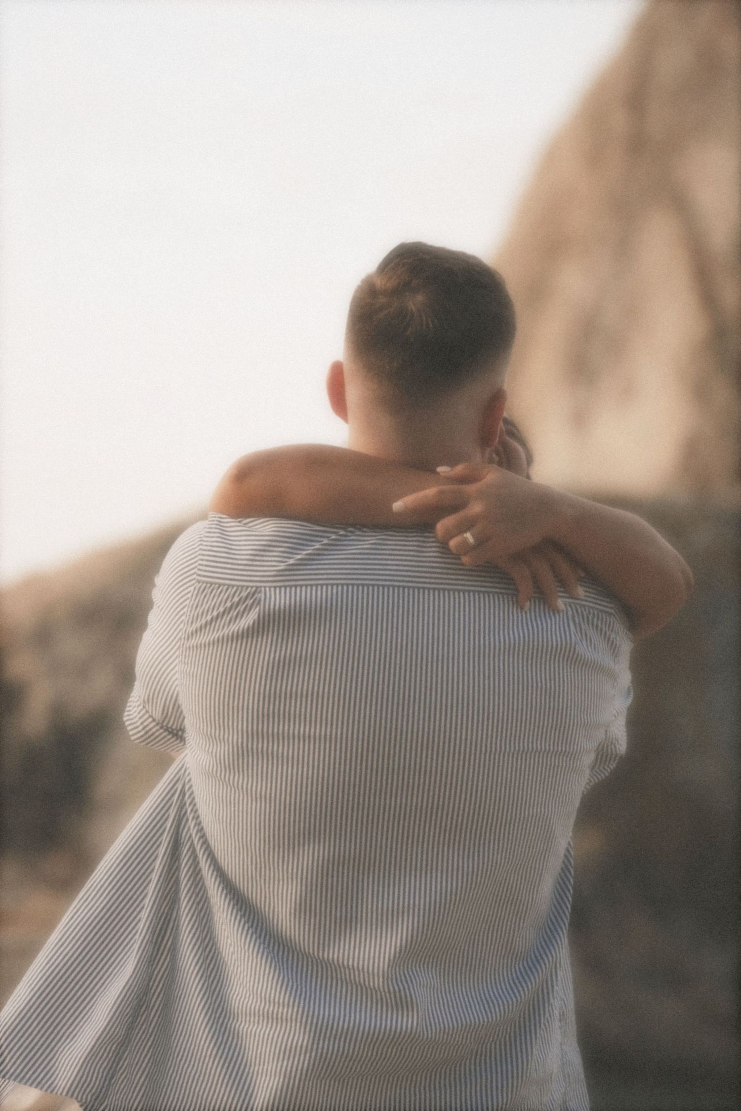 Man hugging a woman, his back to camera, her engagement ring on his shoulder, rocks blurred behind