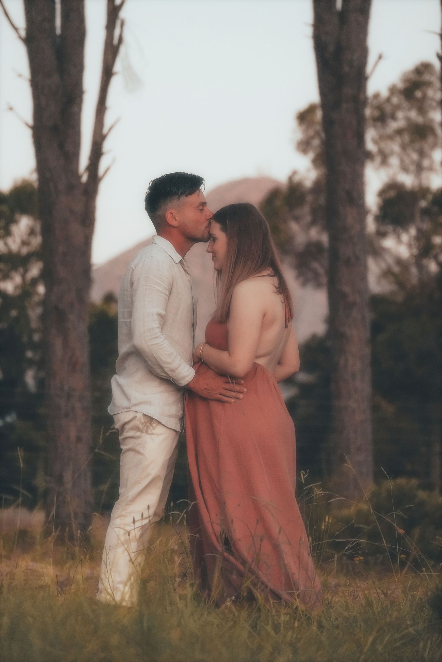 Couple embracing as he kisses her forehead, among tall pines with mountains behind
