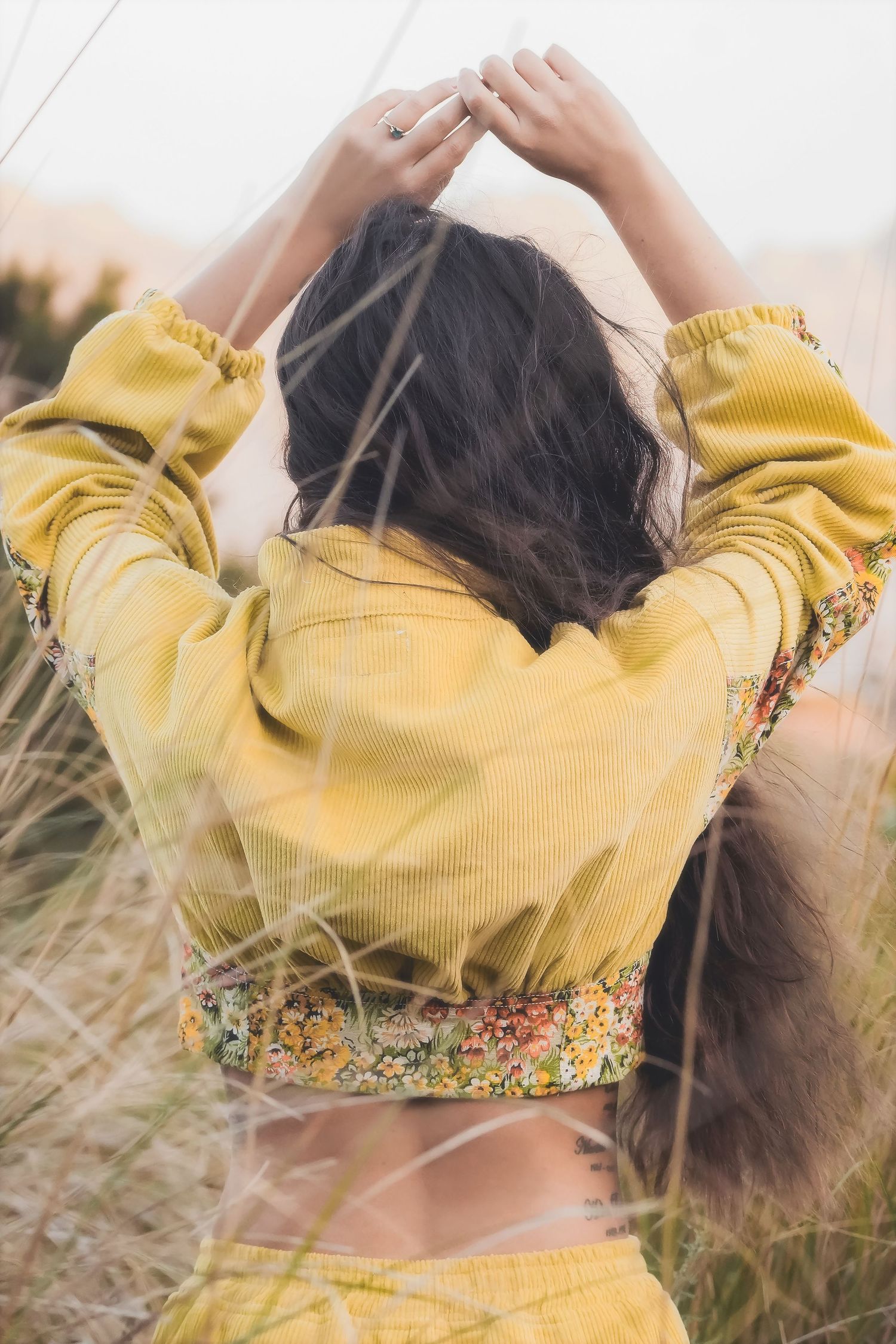 Woman seen from behind with raised arms, long hair falling over a yellow jacket